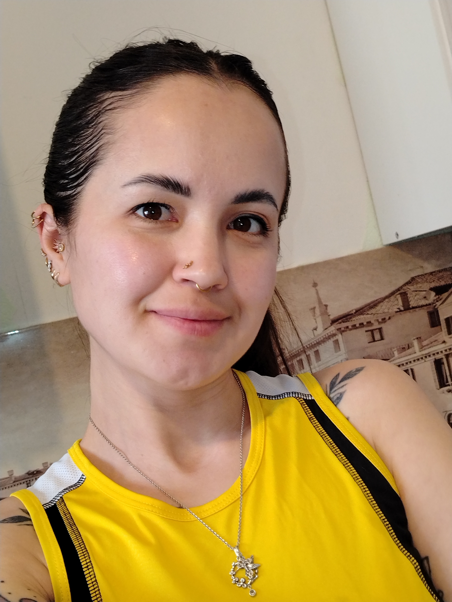A young woman is featured in a close-up, chest-up selfie. Her dark hair is pulled back, appearing wet or slicked, showcasing a distinct hairline with baby hairs. She has brown eyes and a subtle, friendly smile, with clear skin that appears slightly dewy.
Notable details include multiple piercings: a septum ring, two nostril piercings (one stud, one hoop), and numerous ear piercings on her left ear including helix, conch, and lobe piercings. She wears a bright yellow athletic-style tank top with black and white trim along the shoulders and sides. A silver chain necklace with an intricate, ornate pendant (possibly depicting a crown-like design or a stylized insect/flower) is visible around her neck. Tattoos include a leafy branch design on her right shoulder/upper arm and another faint tattoo on her left arm.
The setting appears to be indoors, against a light-colored wall. Behind her, a large, sepia-toned picture or mural depicts classical European-style buildings, featuring arched windows and detailed facades, suggestive of a scene from a city like Venice. The lighting is bright and even, illuminating her face and the details of her attire and jewelry. There is no visible text in the image.