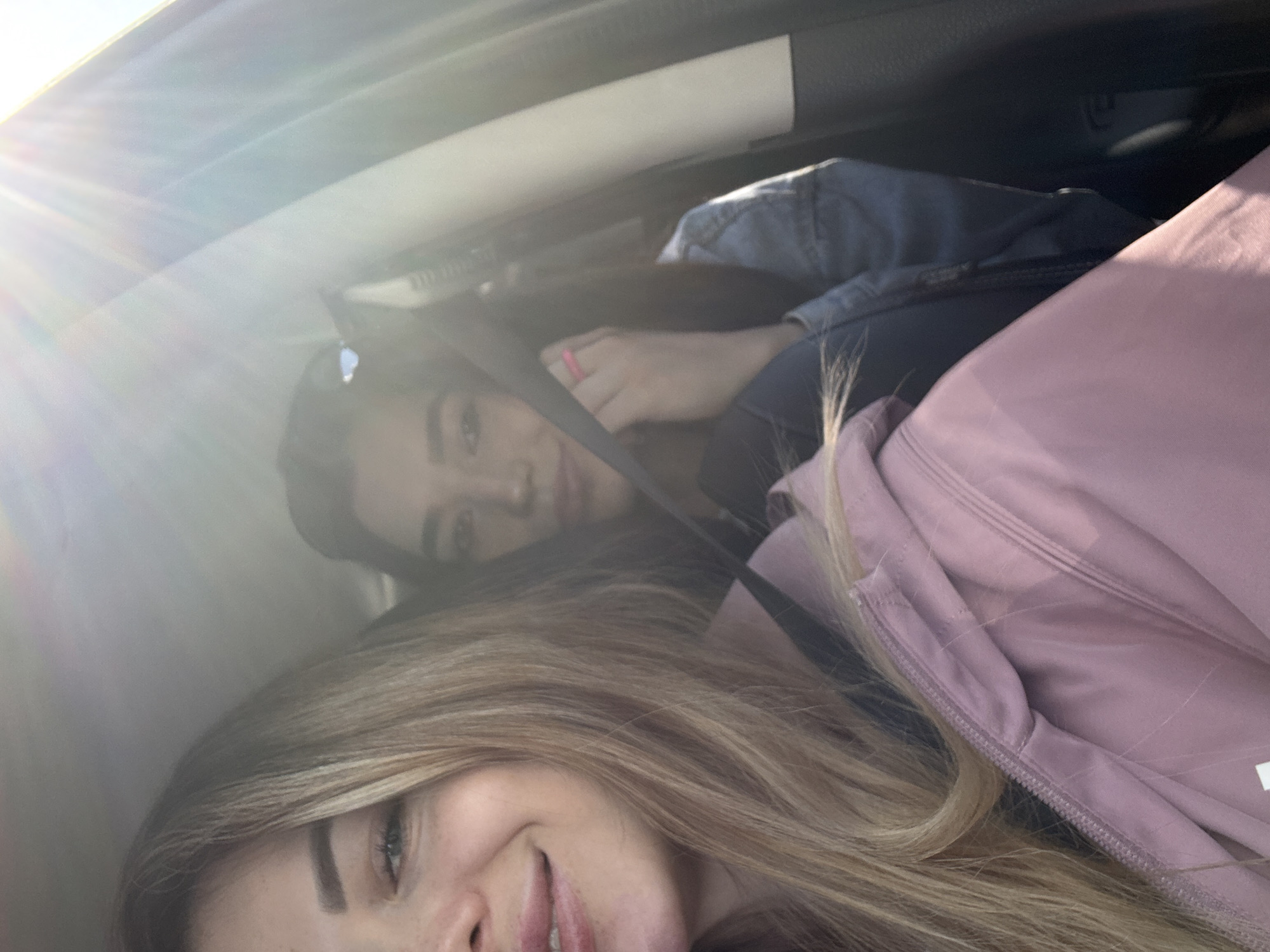 The image captures two women inside what appears to be a car, oriented as if viewed from a tilted perspective. Bright sunlight streams into the vehicle from the upper left, causing a lens flare effect.
In the foreground, the bottom half of a woman's face is visible, featuring light blonde hair, blue eyes, and a slight smile on her lips. She is wearing a soft pink or lavender top or jacket with a visible zipper and textured fabric. Her hair partially covers her face and chest.
Behind and slightly above her, another woman is visible, wearing a light blue denim jacket and a dark seatbelt across her chest. She has dark hair and is looking towards the viewer with a neutral or slightly smirking expression. A bright pink ring is visible on her right hand. The car's interior, including parts of the headliner and window frame, is visible around them.
The scene suggests a casual moment, possibly a selfie taken while traveling or parked. The time of day is clearly daytime, given the strong sun and bright lighting. No specific outdoor location is discernible, and no text is visible in the image. Both women appear relaxed and at ease.