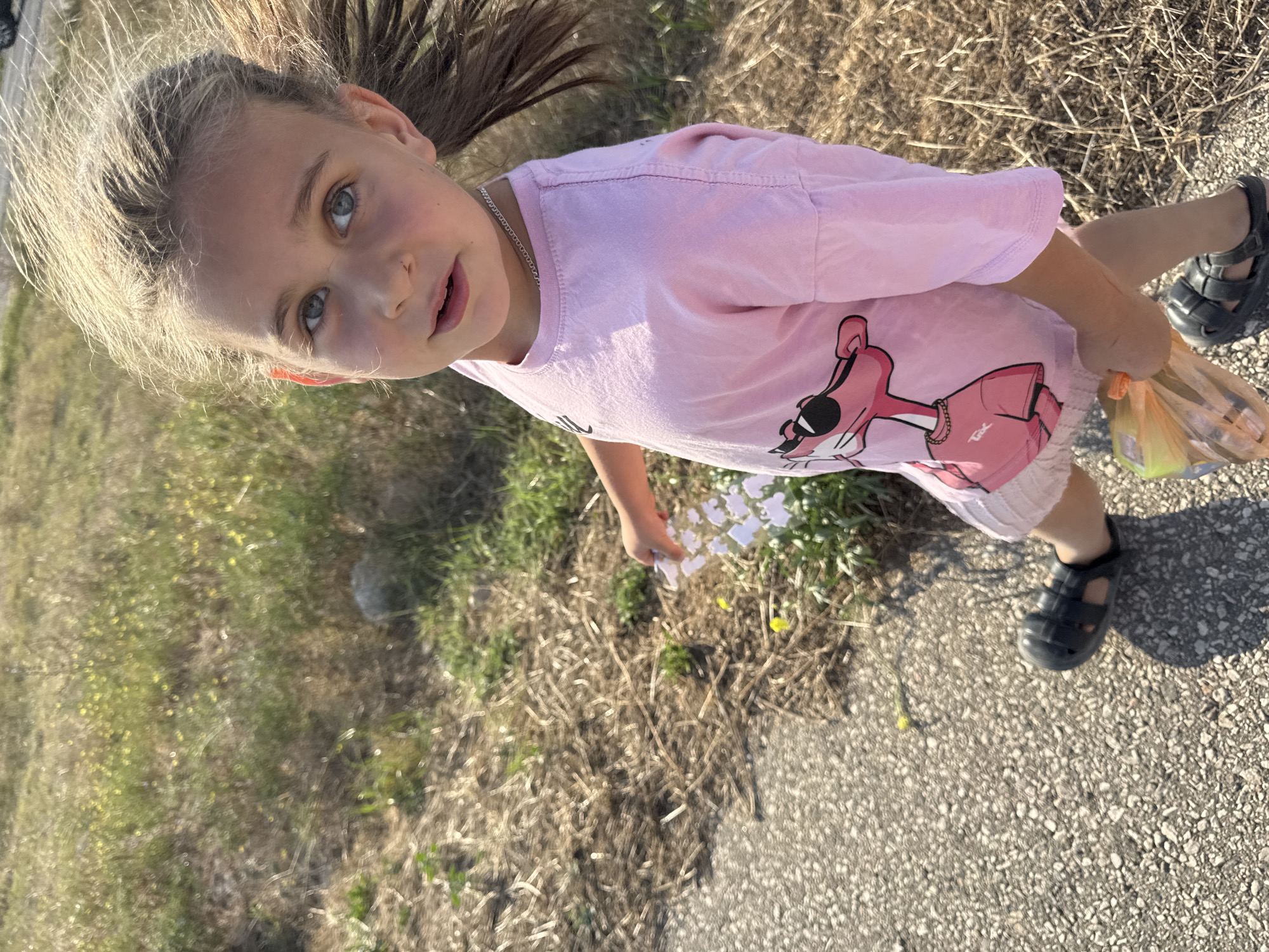 A young girl with blonde hair tied back in a ponytail and blue eyes is walking on a paved path. She is wearing a pink t-shirt with a cartoon character graphic of Pink Panther wearing sunglasses, and light-colored shorts. She has on black sandals and is holding a transparent orange plastic bag in her right hand. The environment appears to be an outdoor setting with dry grass and some green vegetation to her right. The scene is captured from a slightly elevated perspective looking down at the girl. The lighting suggests it is daytime with sunlight casting shadows. The location is identified as Yevpatoriya, Ukraine.