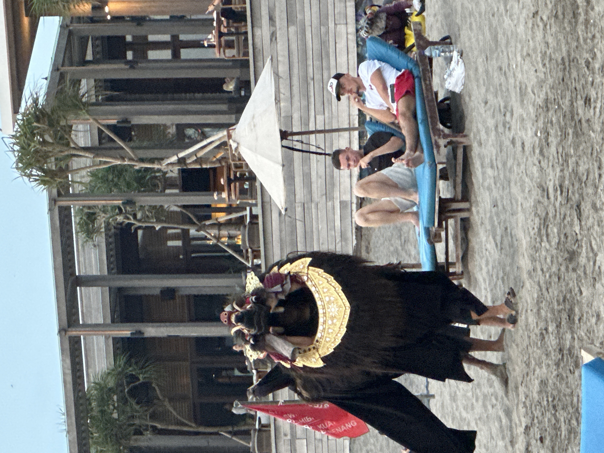 The image depicts a scene on a beach in North Kuta, Indonesia. In the foreground, a person in a traditional Balinese Barong costume, characterized by its elaborate headdress and dark, shaggy fur, stands on the sand. A red flag with white text is planted to the left of the Barong. Further in the background, two individuals are relaxing on blue lounge chairs under a white umbrella. One man is wearing a black t-shirt and white shorts, while the other wears a white t-shirt, red shorts, and a baseball cap. A bottle of water is visible on the sand near the lounge chairs. The backdrop features a multi-story building with wooden architectural elements, possibly a restaurant or resort, with tables and chairs set up on elevated platforms. The environment is sandy with sparse vegetation, indicative of a beach setting. The lighting suggests it might be late afternoon or early evening.