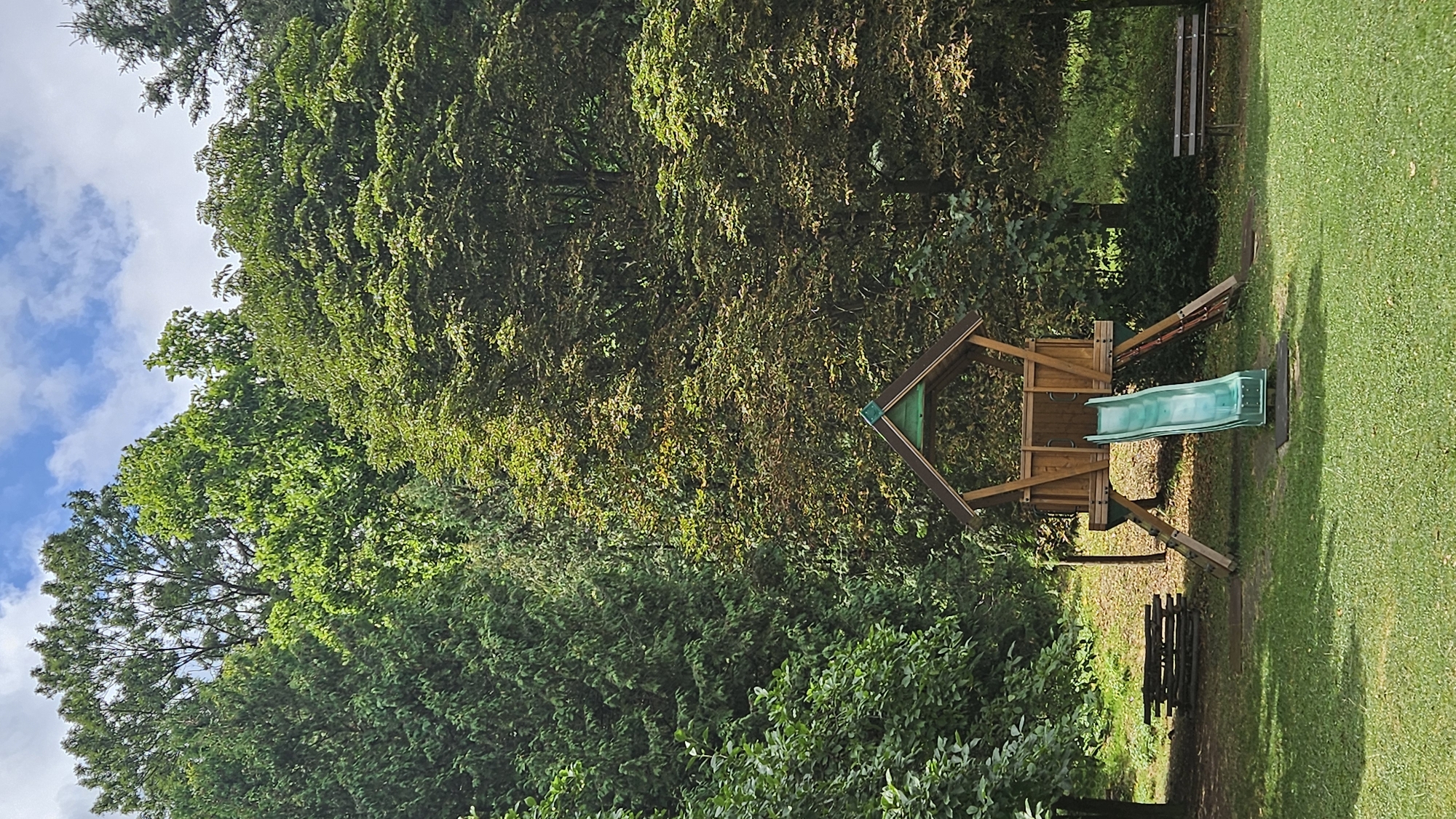 The image captures a natural outdoor setting, likely a park or recreational area. Dominating the frame is a wooden play structure with a green slide, situated on a grassy lawn. The playhouse has a gabled roof and wooden railings. To the left of the play structure, a wooden bench is visible. To the right, another park bench is positioned under a canopy of trees. The background is filled with lush, dense foliage from a variety of trees, suggesting a forested or heavily wooded environment. The sky above is partly cloudy, with patches of blue visible. The overall impression is one of a serene, natural landscape suitable for outdoor activities. This scene is located in the city of Prešov, Slovakia, specifically within the Šariš National Park, known for its natural beauty and recreational opportunities.
