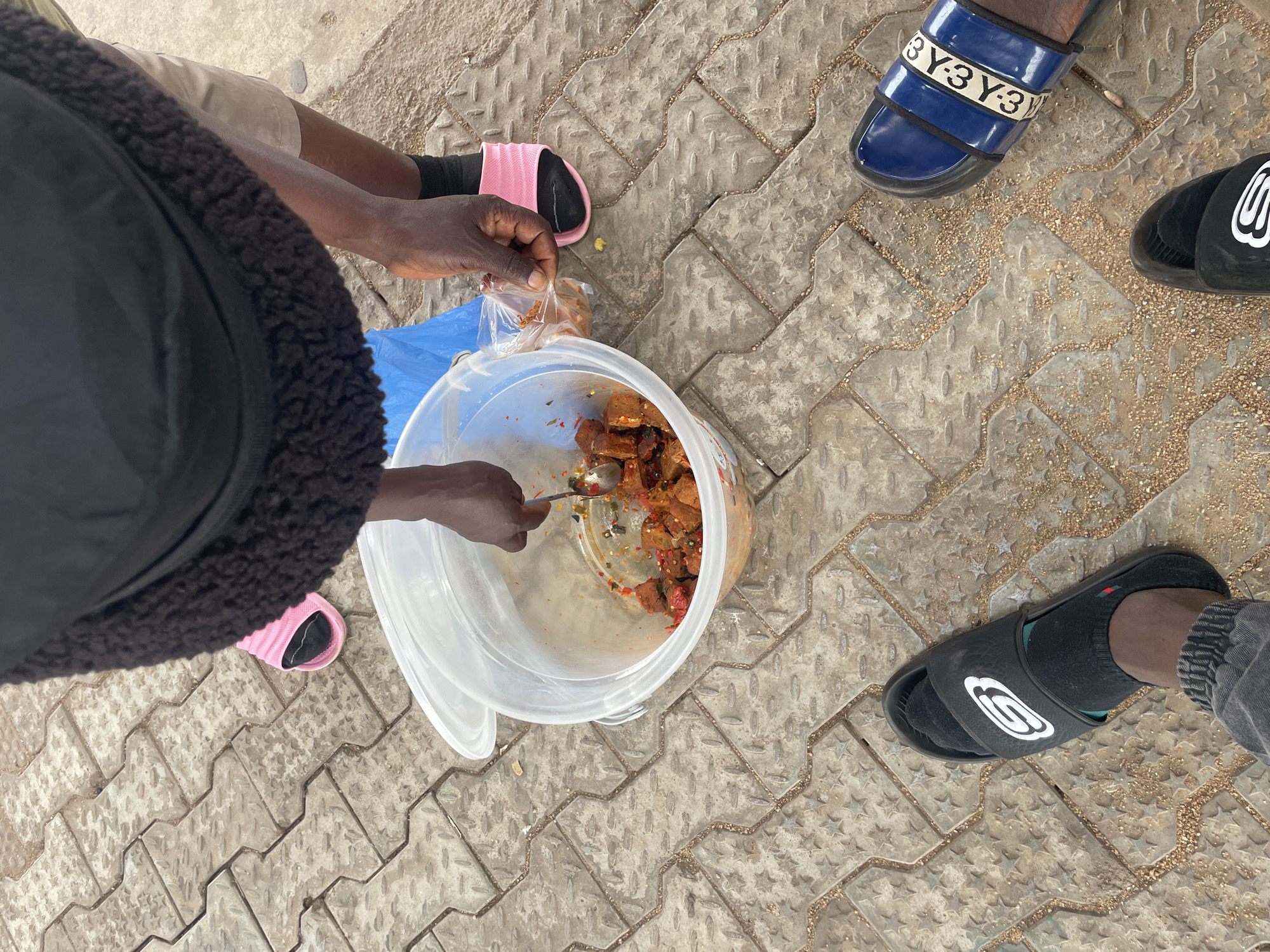 A top-down medium shot shows a person wearing a black jacket with a fur collar standing on a patterned concrete surface. The individual is using a spoon to scoop food from a large, translucent plastic bucket into a small, clear plastic bag. The food appears to be diced and seasoned with various spices. Several pairs of feet in sandals are positioned around the bucket on the same concrete surface. The location is indicated as Jos, Nigeria, though this is not directly visible in the image but is inferred from provided context.