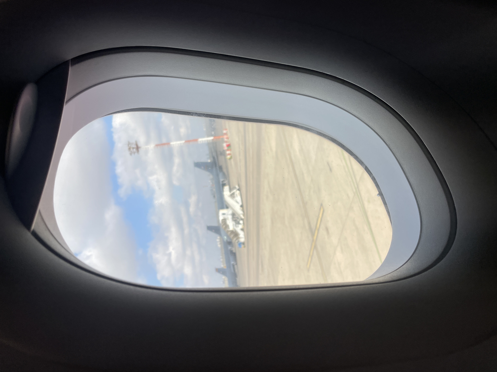 The image shows an airplane window with a view of an airport tarmac on a partly cloudy day. In the distance, two airplanes are visible, along with a set of mobile stairs parked near one of them. A tall, red and white striped tower, likely an airport marker or antenna, stands prominently on the right side of the frame. The tarmac is a paved surface with yellow markings. The foreground is dominated by the dark interior of the airplane's cabin, framing the exterior view. The location is identified as the city of Yagel, Israel.