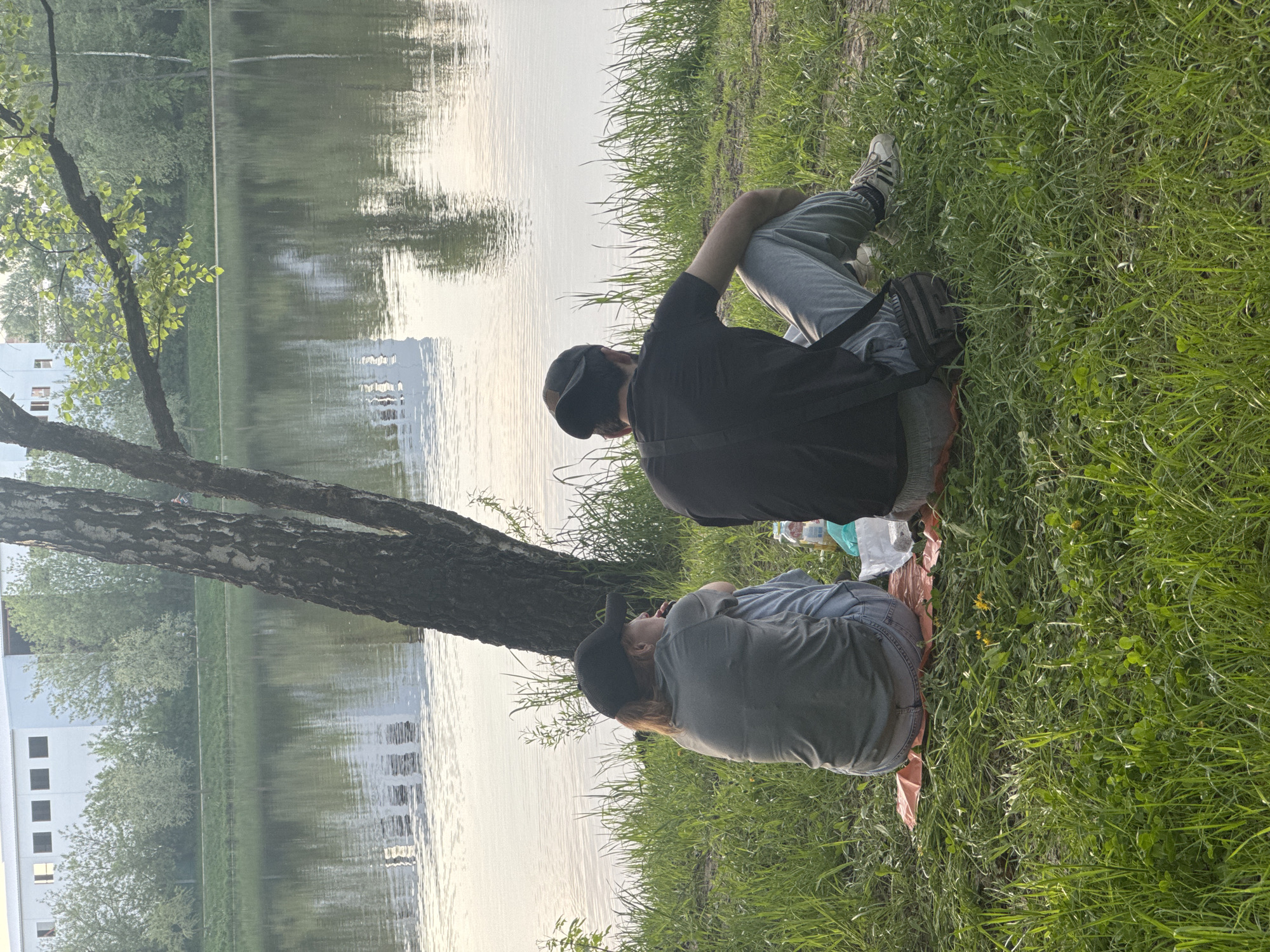 Two individuals are sitting on the grass near a body of water. A tree trunk is situated close to the water's edge, adjacent to the individuals. The water reflects the surrounding buildings and sky. The scene is likely taking place in a park or recreational area in Malakhovka, Russia, as per the user's instructions, although this cannot be definitively determined from the image alone.
One person is wearing a black shirt and a cap. The other person is wearing a gray shirt and a cap. They are sitting on a blanket. A bag and some bottles are on the blanket between them.
The environment consists of grass, water, trees, and buildings in the background.