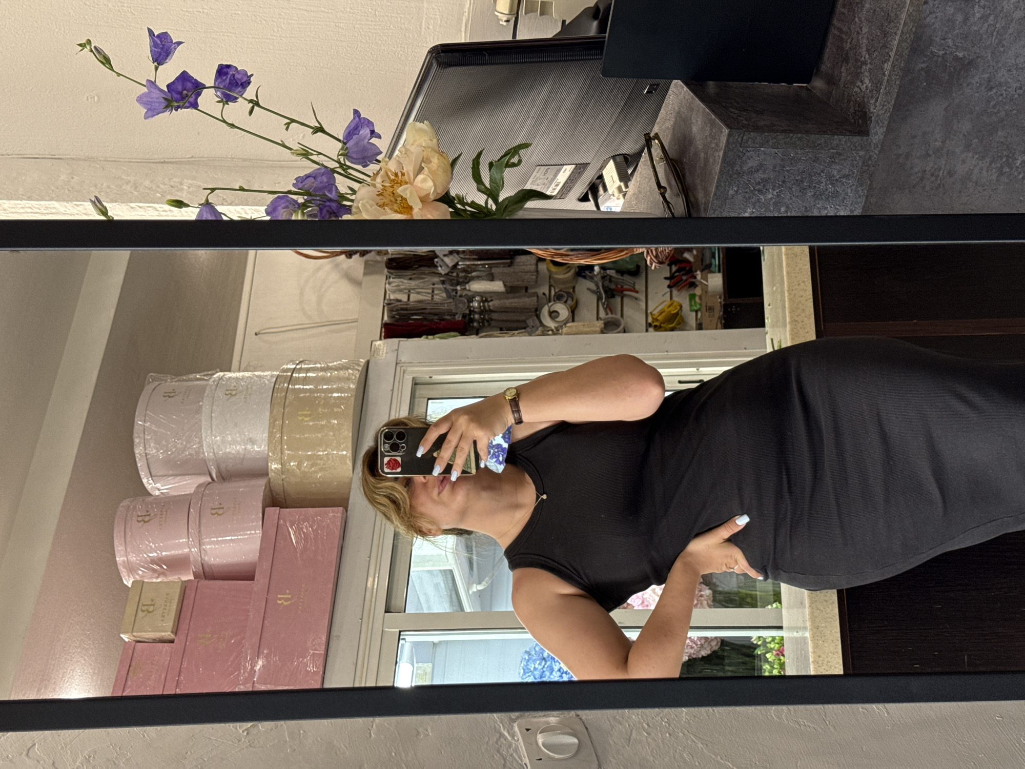 The image shows a young woman standing in a shop and taking a selfie in a large mirror. She is wearing a black sleeveless dress and has light blue nail polish. She is wearing a delicate necklace and a wristwatch. Behind her, on shelves, are stacked pink and white boxes, some circular and some rectangular, with a "B" logo on them. To the right of the woman, a vase of flowers, including purple bellflowers and a light pink peony, sits on a desk next to a computer monitor. The background appears to be a retail space with various items on display, including what look like decorative tassels and tools. The woman's reflection captures her from the chest up, with her hand on her hip. The scene is likely located in Vladikavkaz, Russia, given the context of the request, though no explicit visual cues within the image confirm this.