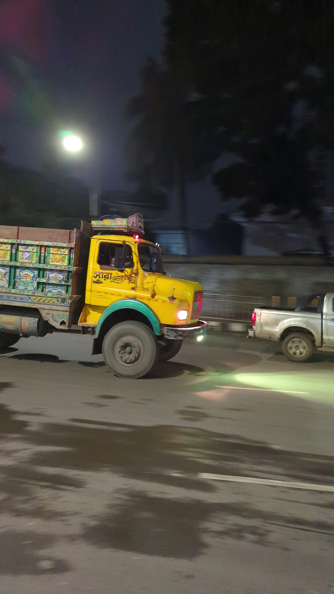 The image depicts a night scene on a city street in Dhaka, Bangladesh. A yellow truck with colorful decorative paintings on its side is prominently featured, traveling on the wet road. The truck has a large tire and its headlights are illuminated. To the right of the truck, a silver pickup truck is also visible, its headlights shining on the road. The street surface is wet, reflecting the lights from the vehicles and streetlights, with dark shadows of trees visible in the background. A bright street light illuminates the upper left portion of the image. The overall impression is one of urban transportation at night, with the wet conditions suggesting recent rain.