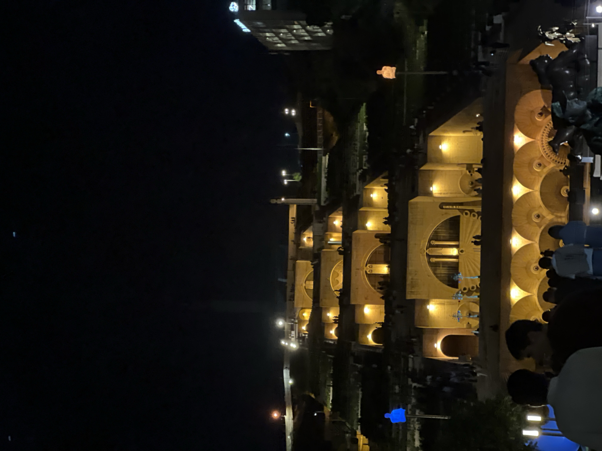 The image depicts the Yerevan Cascade in Yerevan, Armenia, at night. The Cascade, a monumental stairway with fountains and sculptures, is illuminated, creating a warm glow. People are seen walking along the steps and gathering near the fountains. The scene also captures the surrounding cityscape, including a multi-story building and street lighting.