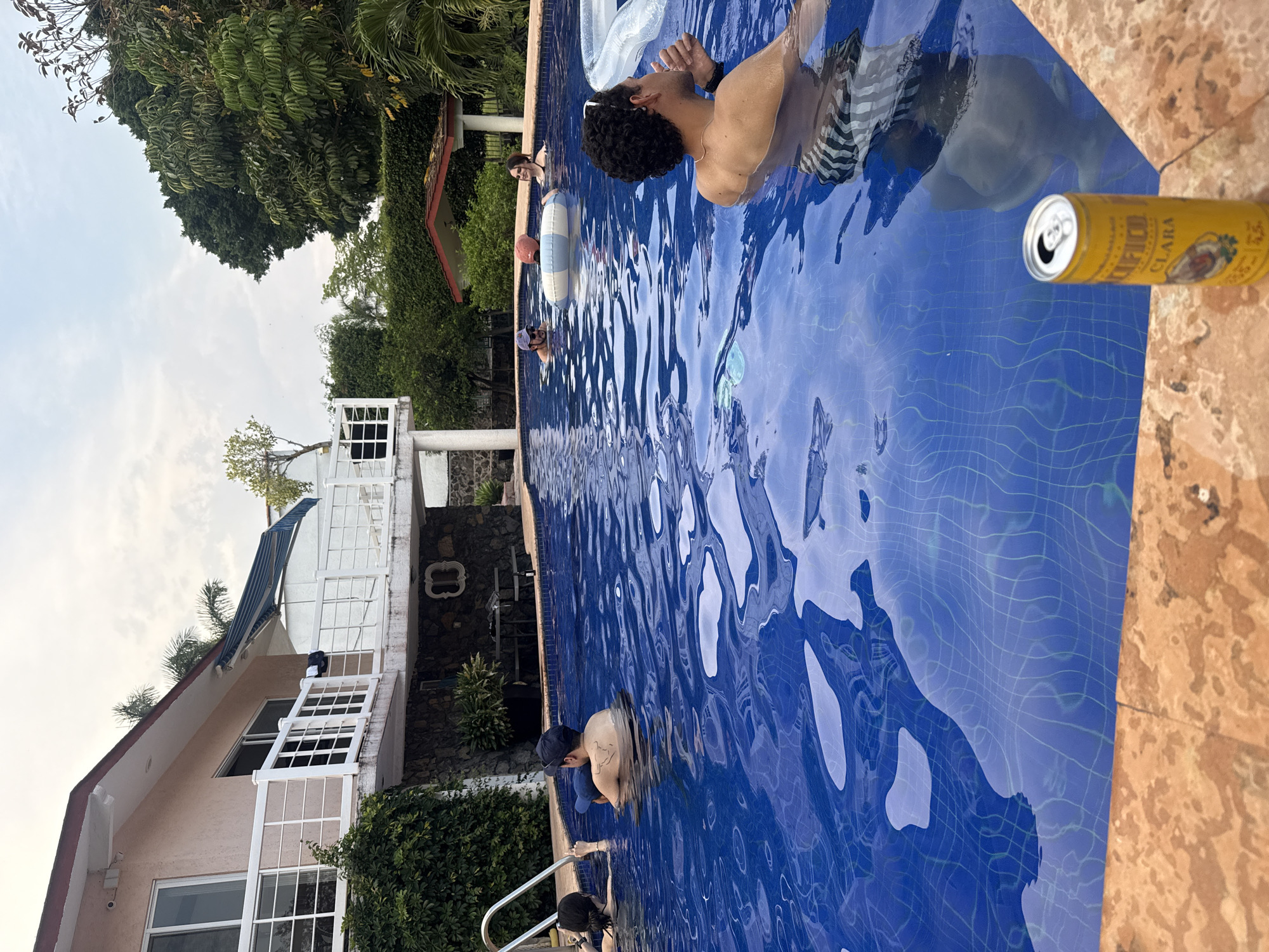 The image depicts a swimming pool scene in Oaxtepec, Mexico. The pool is blue-tiled and contains several people, including a man with curly hair who is swimming, and another individual wearing a baseball cap. A partially visible beer can sits on the pool's edge. Behind the pool is a two-story house with white balconies and a stone wall feature. Lush green trees and other vegetation surround the area. The sky is partly cloudy.