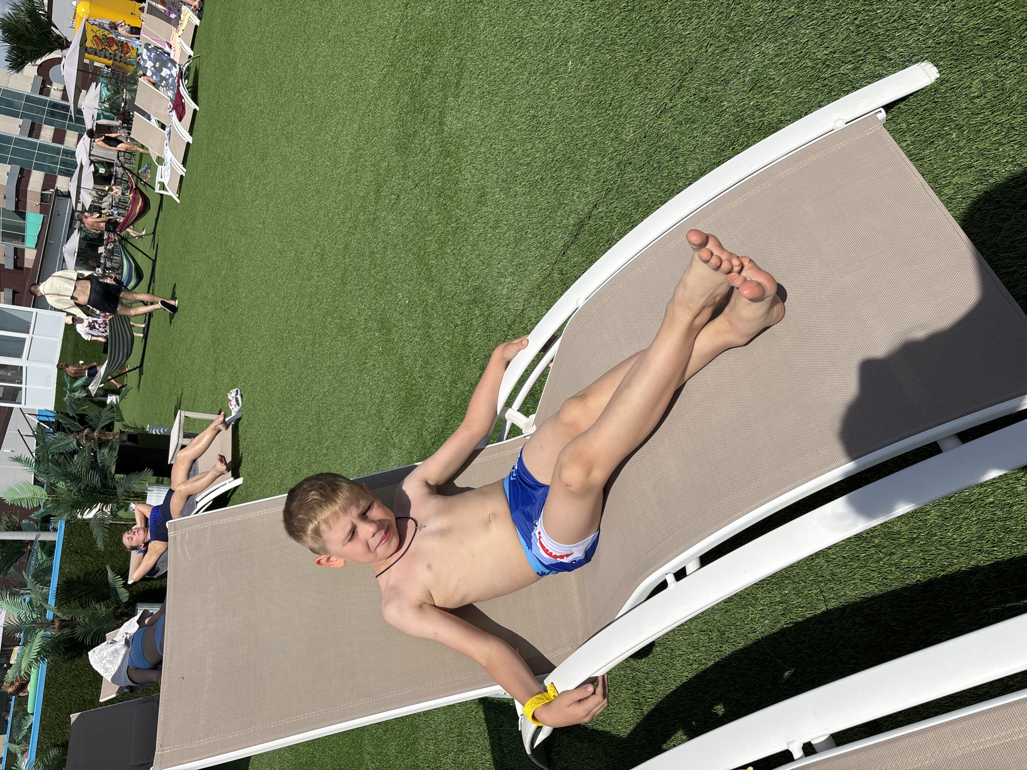 A young boy is sunbathing on a lounge chair on a grassy area, possibly a resort or park. He is wearing blue swim trunks with a shark pattern and a black necklace. His left arm is resting on the armrest of the chair, and he is wearing a yellow wristband on his right wrist. His eyes are closed, and he appears relaxed, enjoying the sunny weather. In the background, other people are visible lounging on chairs, and palm trees are scattered throughout the area, suggesting a warm climate. The setting appears to be outdoors, with a clear blue sky and bright sunlight, indicating it's daytime. The overall mood is one of leisure and relaxation. The cropped images show close-ups of the boy's feet, the lounge chair, and the grassy ground. There is no visible text in the image.