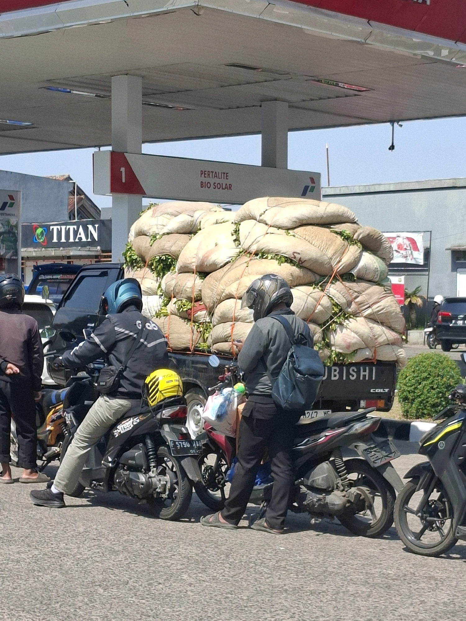 The image depicts a sunny outdoor scene at a gas station in Cianjur, Indonesia. A black Mitsubishi Colt pickup truck is parked and heavily laden with large, burlap sacks filled with green produce, possibly tea leaves, tied down with ropes. Several motorcycles are also present. In the foreground, a person wearing a black jacket with a blue helmet and a black bag over their shoulder is seated on a motorcycle. To their left, another person in a dark top and black pants stands near their motorcycle. Behind them, a black pickup truck with a driver in a dark jacket and helmet is visible. The gas station features a canopy with signage indicating "PERTALITE BIO SOLAR" and a prominent red and white number "1". In the background, a gray building and a lush green bush are visible. The ground is paved with asphalt.