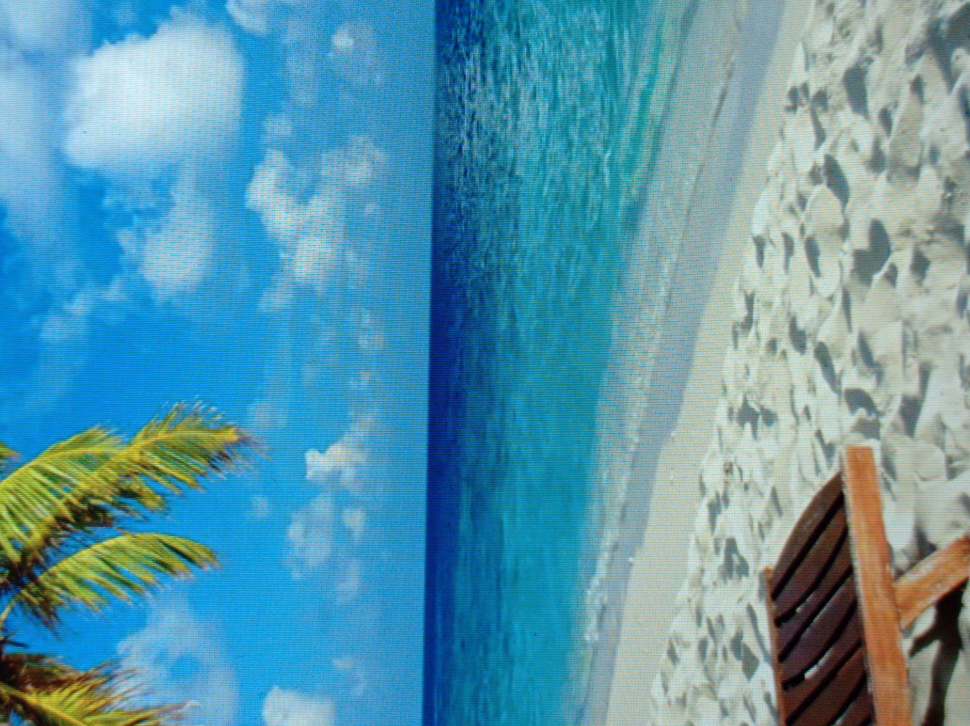 The image depicts a serene tropical beach scene. A wooden lounge chair is positioned on white sand, facing a clear turquoise ocean under a bright blue sky dotted with fluffy white clouds. Palm fronds from a nearby tree overhang the top left corner of the frame, adding to the tropical ambiance. The waves gently lap at the shore, creating a peaceful and inviting atmosphere. There are no people or buildings visible in the image, suggesting a secluded and tranquil location. The scene evokes a sense of relaxation and escape. The image does not contain any elements that would suggest a connection to the city of Mukachevo, Ukraine.