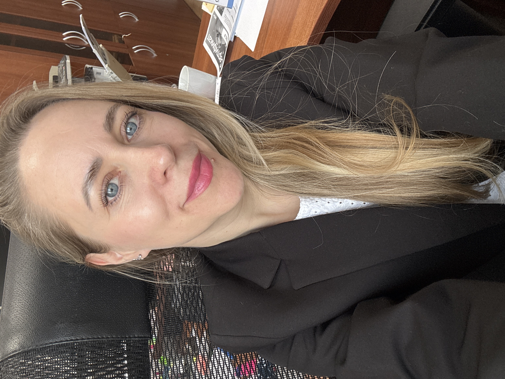 A light-skinned woman with blonde hair, blue eyes, and pink lipstick is taking a selfie. She is wearing a black blazer over what appears to be a white shirt. She is seated in a black office chair. The background includes a wooden desk with papers and a mug, along with a wooden cabinet with metallic handles.
There is no information in the image to support the claim that the location is in the city of Chaikovsky, Russia.