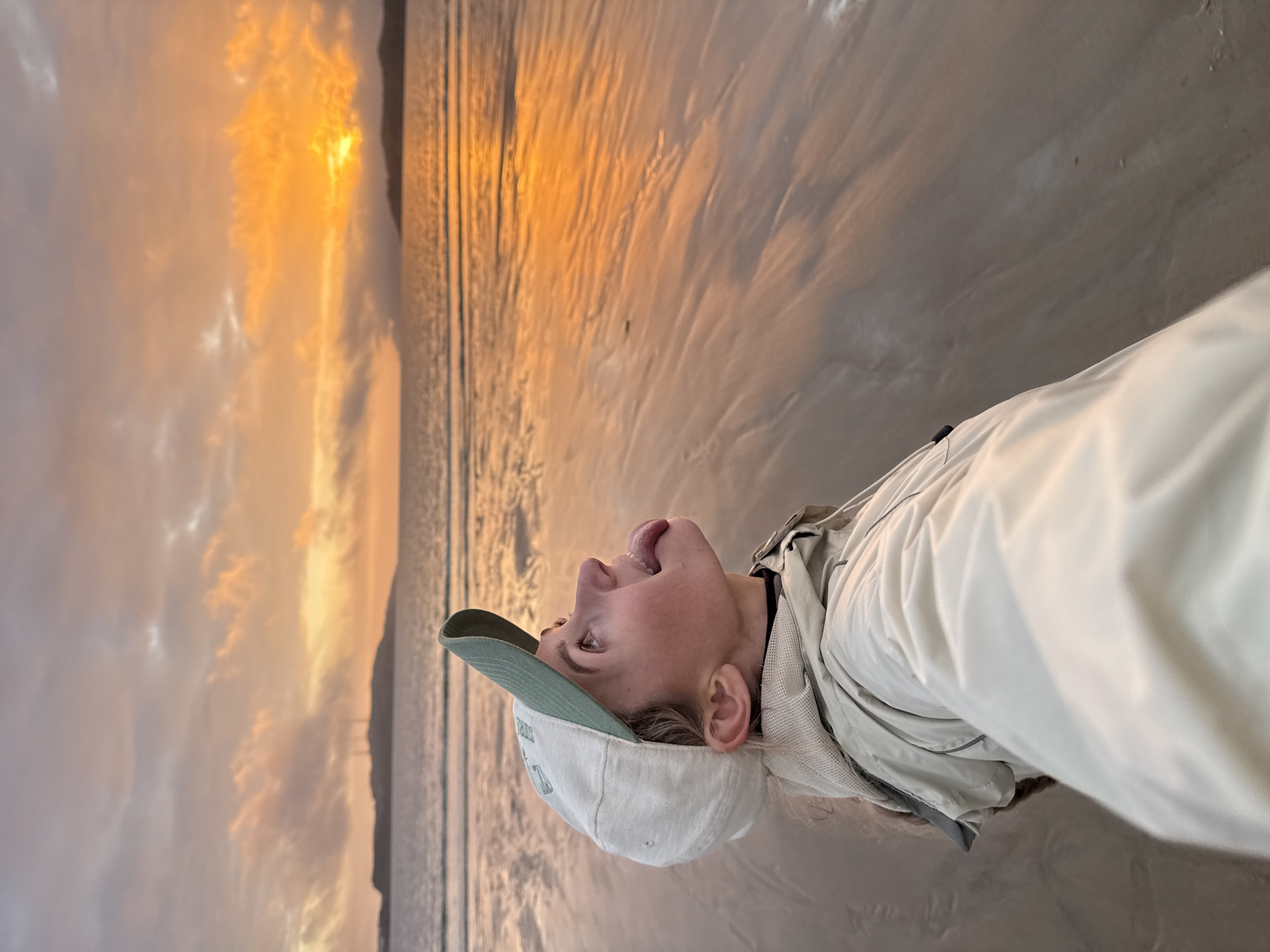 In Teriberka, Russia, a young woman wearing a baseball cap and a light-colored jacket stands on a beach during sunset. The sky is filled with vibrant orange and yellow hues, with clouds casting dramatic shadows. The ocean stretches out before her, reflecting the colors of the sky, and the wet sand on the beach also shows these reflections. The woman is looking up towards the sky with her tongue sticking out, appearing playful. The overall scene captures a moment of joy and appreciation for the natural beauty of the sunset over the sea.
