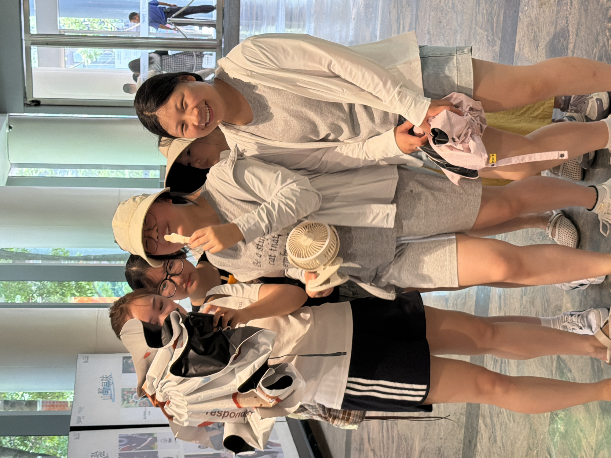 This is a medium shot of five young Asian women and girls standing in an indoor public space. The woman in the foreground on the right is smiling and looking at the camera. She is wearing a grey t-shirt and a white open cardigan, with light blue denim shorts. To her left, a girl in a light-colored hat is holding a small, white fan and eating. She is wearing a grey long-sleeved shirt and grey shorts. To her left, a woman is holding a black smartphone and looking at it. She is wearing a white t-shirt with black stripes on the hem of her skirt. To her left, another girl with glasses is looking at the woman with the phone. Another girl's face is partially visible behind her. In the background, a glass door leads to an outdoor area where two men are visible. The floor appears to be tiled, and the walls are light-colored, with some posters or displays on them. The environment suggests a museum, exhibition hall, or a similar public building. The overall impression is of a group of friends enjoying an outing.