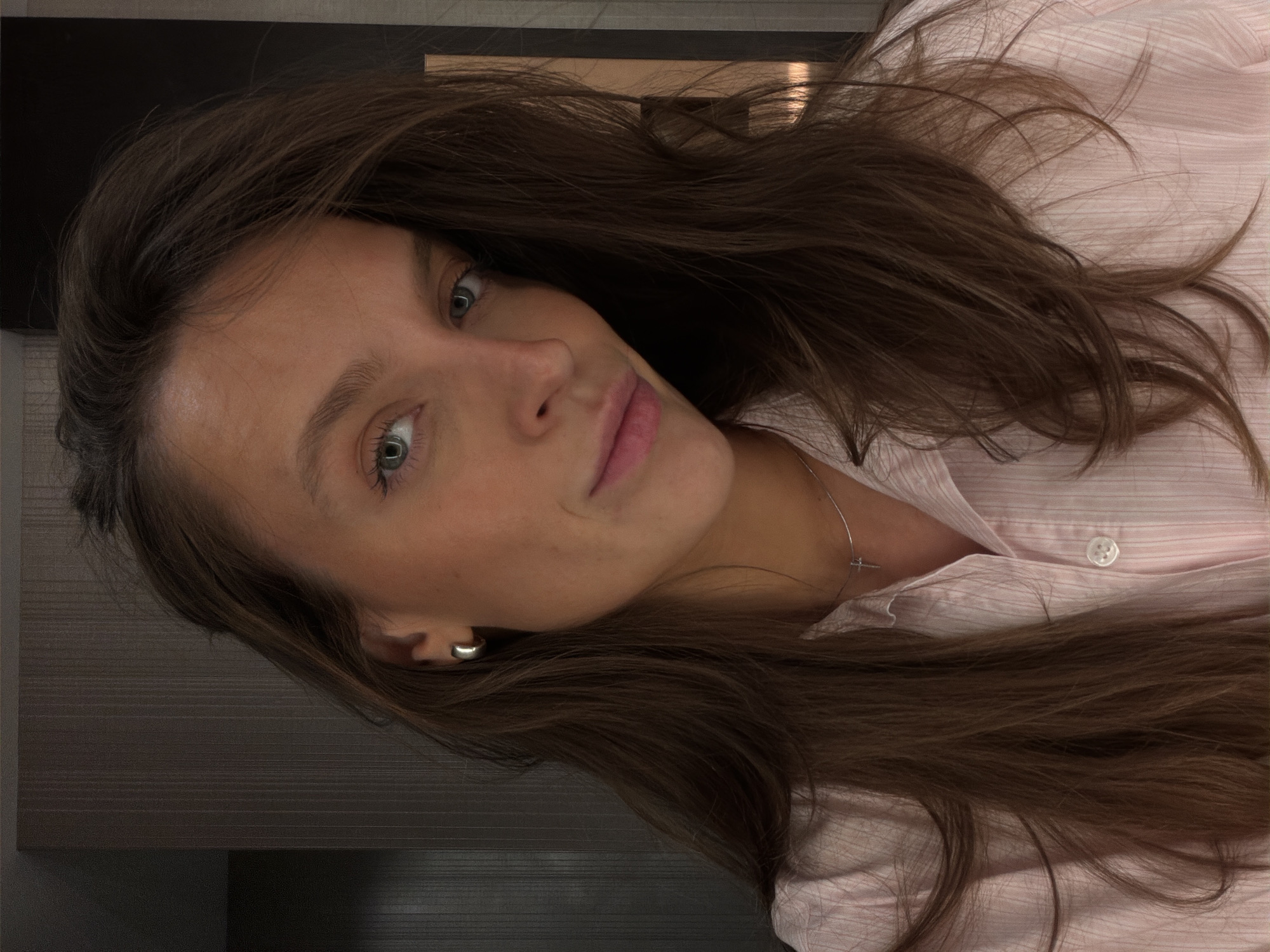 The image shows a close-up, medium shot of a young woman with long, brown hair, wearing a light pink striped button-down shirt and a silver cross necklace. She has blue eyes and light skin with a few freckles. She is wearing a silver hoop earring in her left ear. The background appears to be an indoor setting with a dark, textured wall and a hint of a lighter-colored wall on the right. No specific location in Budva, Montenegro is identifiable from the image. The woman is looking towards the camera with a slight smile.