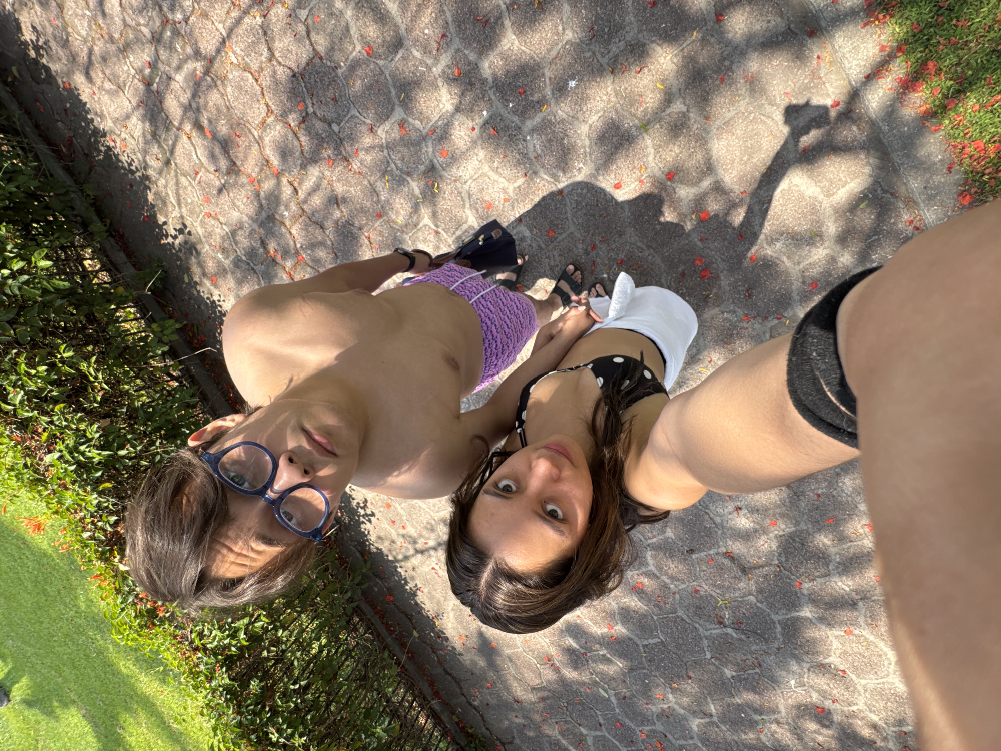 A point-of-view medium shot depicts a light-skinned man and woman in Cocoyoc, Mexico. The man is wearing glasses and purple bathing trunks. The woman has brown hair and a black and white polka dot bathing suit. Both are standing on a walkway with red flowers and leaves scattered across its surface. Bushes line the left side of the walkway. The shadow of a hand holding a smartphone is visible.