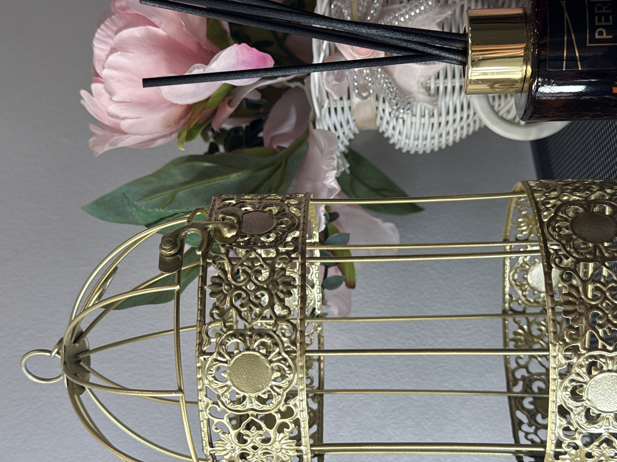 The image showcases a decorative arrangement featuring a golden, ornate birdcage, artificial flowers, and a reed diffuser. In the foreground, positioned to the right, is a golden metal decorative cage or birdcage. It exhibits intricate filigree patterns on its vertical supports and simple horizontal bars, with a detailed latch mechanism visible at the top.
To the left of the cage, a bouquet of artificial pink flowers, likely peonies, with vibrant green leaves, fills the mid-ground. Above and slightly behind the flowers, a dark amber-colored reed diffuser bottle with a shiny golden cap and several black reeds extends upwards. A portion of a white woven decorative basket is visible behind the diffuser.
The background is a plain, light-colored surface, providing a simple backdrop. The lighting is soft and even, highlighting the textures of the metalwork, fabric petals, and the glossy diffuser bottle. On the diffuser bottle, the text "PER" is partially visible, suggesting "PERFUME" or a brand name. No people are present, and the scene is a static display of home decor items, possibly on a shelf or table. There are no specific visual cues within the image to indicate the location as Chaikovsky, Russia.