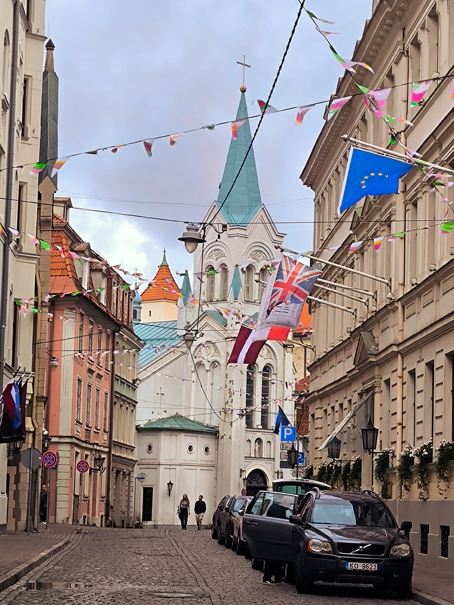 The image shows a narrow, cobblestone street in Riga, Latvia, lined with historic buildings. Colorful flags and bunting are strung across the street, suggesting a festive occasion. Prominently featured is the facade of a white church with a green steeple, topped by a cross. In the foreground, several parked cars are visible, including a dark SUV with its driver's side door open. Two people are walking away from the camera down the street, and another person is partially visible near the entrance of a building on the left. The architecture of the buildings, with their ornate facades and tiled roofs, is characteristic of the Old Town of Riga. The sky is overcast, and the lighting suggests it is daytime, likely overcast or late afternoon.