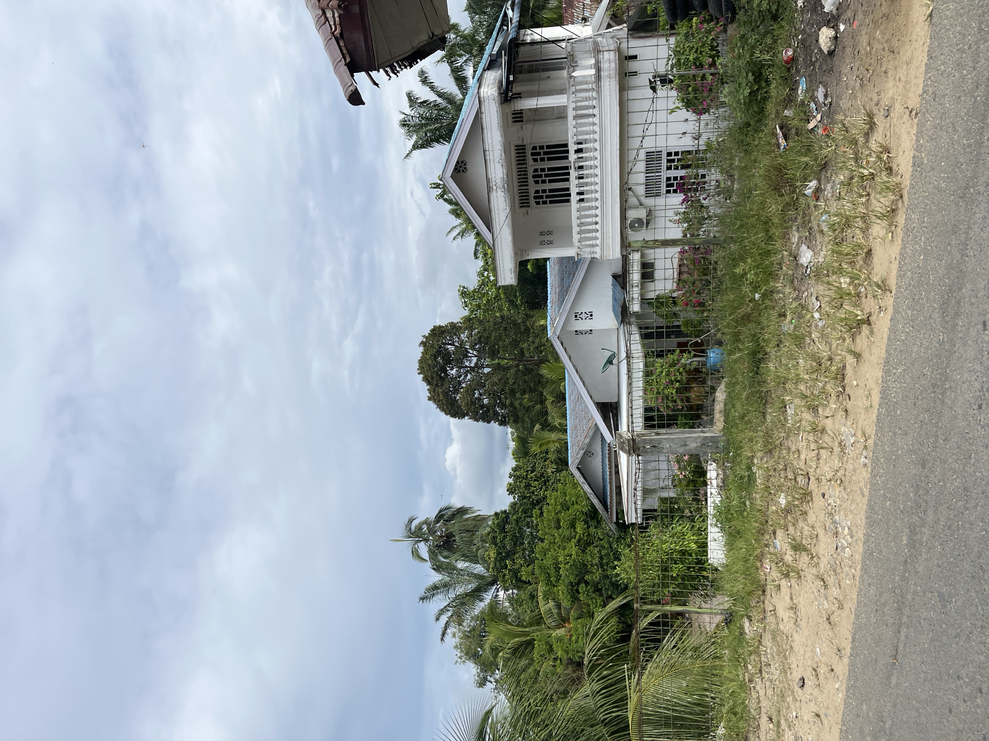 The image depicts a residential street scene in Sukajadi, Indonesia, characterized by two white houses. The house on the right is a two-story structure with a balcony featuring ornate railings and white columns. It has multiple windows on both levels, some with decorative grille work. The house on the left is a single-story dwelling with a similar white exterior and a light blue roof. Both houses are surrounded by lush tropical vegetation, including palm trees and dense foliage, indicating a humid climate. A wire fence separates the properties from the unpaved roadside, which is covered in grass and scattered debris. The sky is overcast, with a mix of light and dark clouds. The overall impression is of a tropical urban environment.