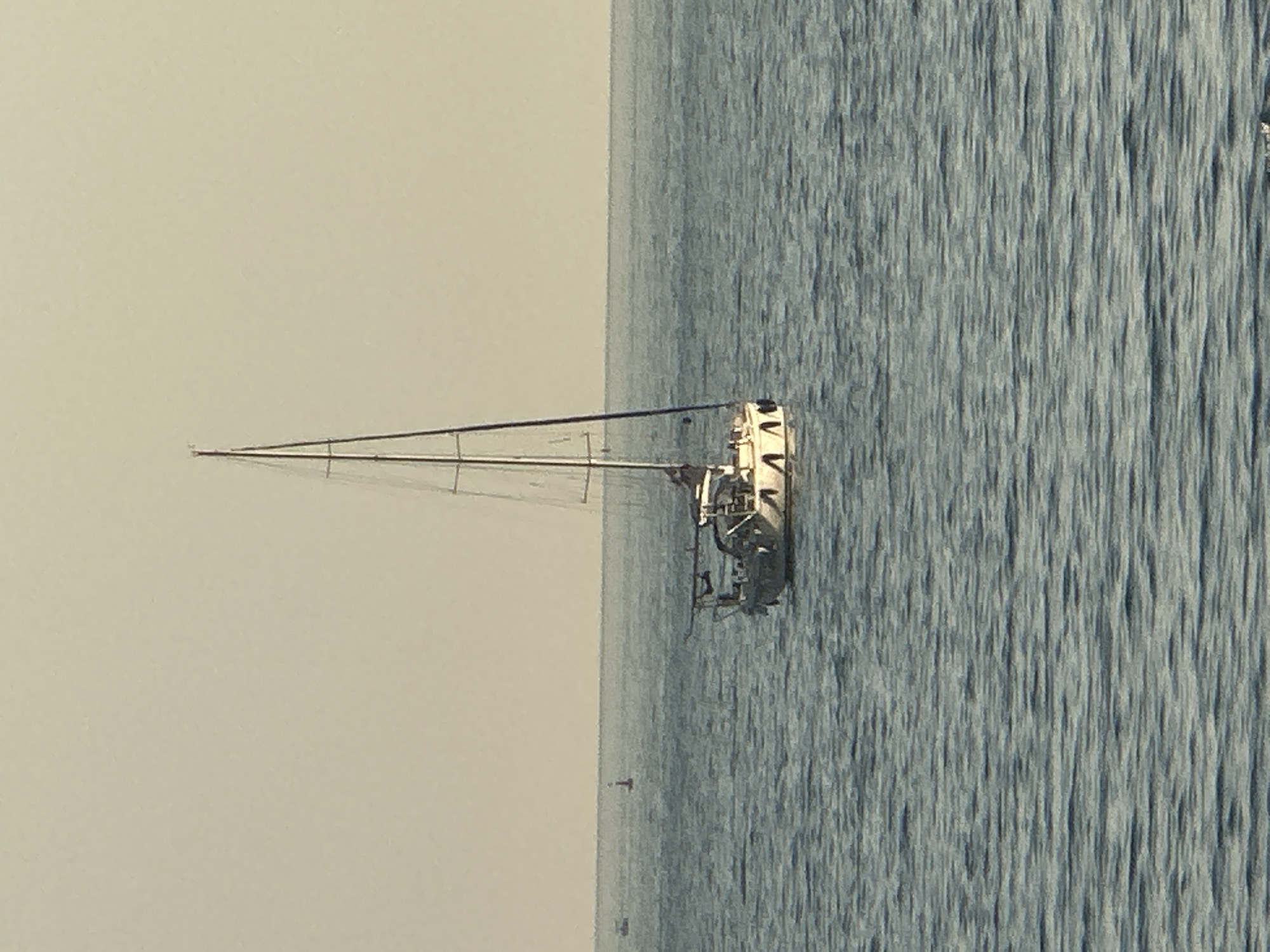 A single-masted sailboat is at anchor in calm, blue water under a pale, hazy sky. The boat, predominantly white with some dark accents, has a covered cockpit area and a visible mast with rigging. In the distance, two faint objects, possibly buoys or other vessels, are visible on the horizon. The foreground is filled with the rippled surface of the sea. The scene is serene and appears to be an open body of water, likely the Adriatic Sea off the coast of Budva, Montenegro, given the context. No people are clearly discernible on the sailboat.