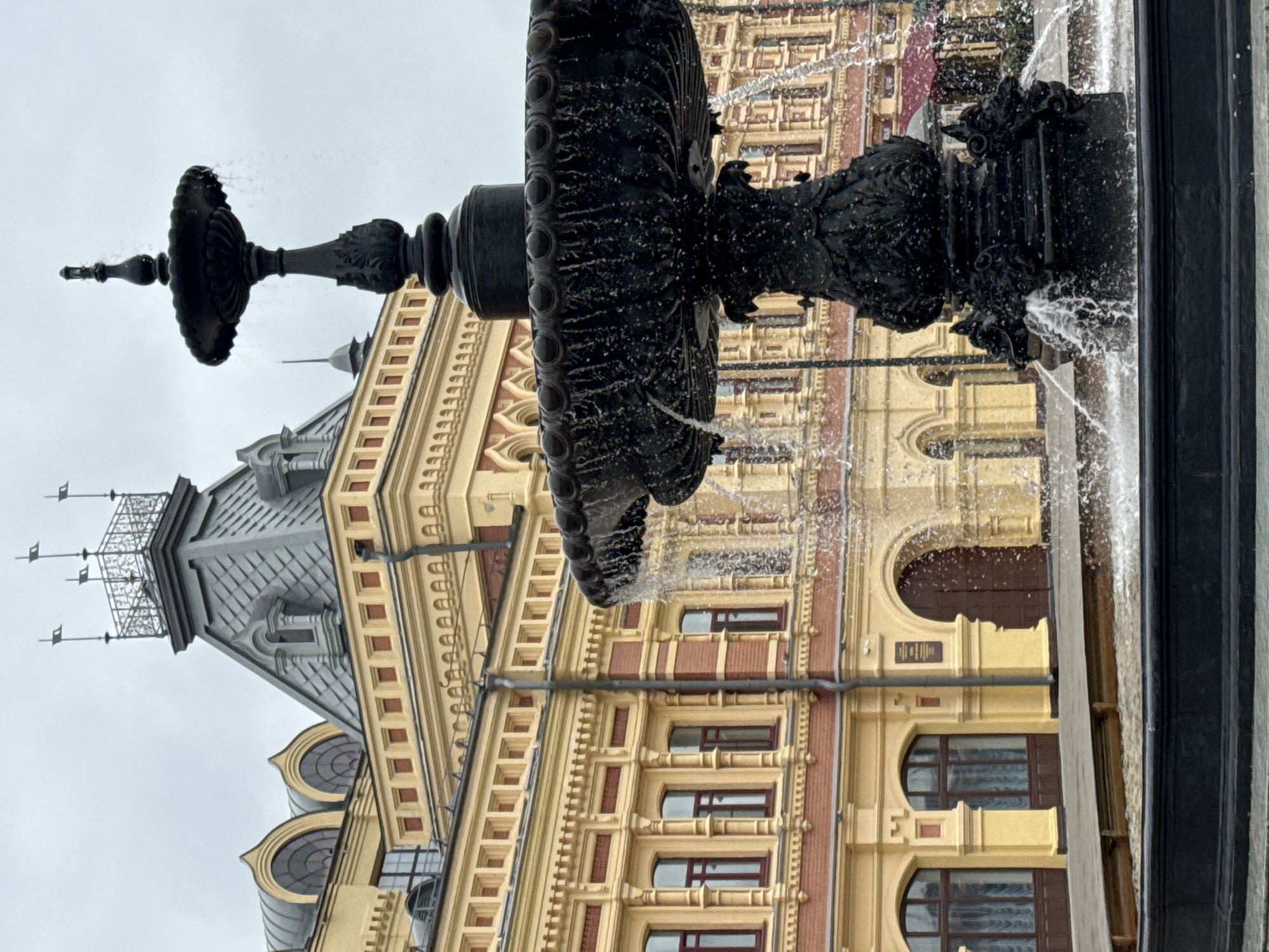 A black ornate fountain with water spraying is in the foreground. Behind the fountain is a large, yellow building with red trim and arched windows and doorways. The sky is overcast. The building is the Telegraph House, located in Nizhny Novgorod, Russia.