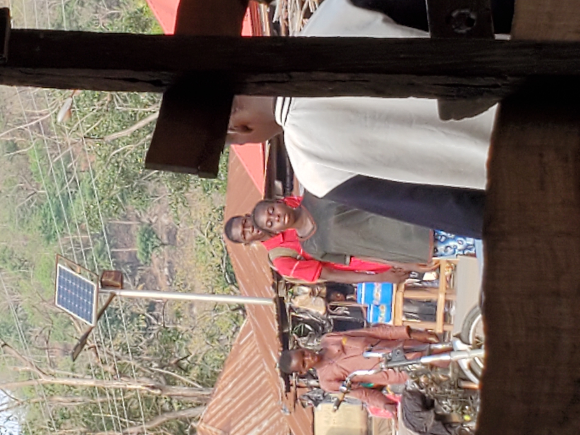 The image shows an outdoor scene in Mararaba, Nigeria. Several people are present: one wears a white shirt with black trim, another a grey shirt, one a red shirt, and another a brown shirt. They appear to be situated near a building with a corrugated metal roof. In the background, there is a solar panel mounted on a pole. A motorcycle is also visible in the scene. The environment includes trees and potentially other buildings. The view is partly obstructed by wooden beams in the foreground.