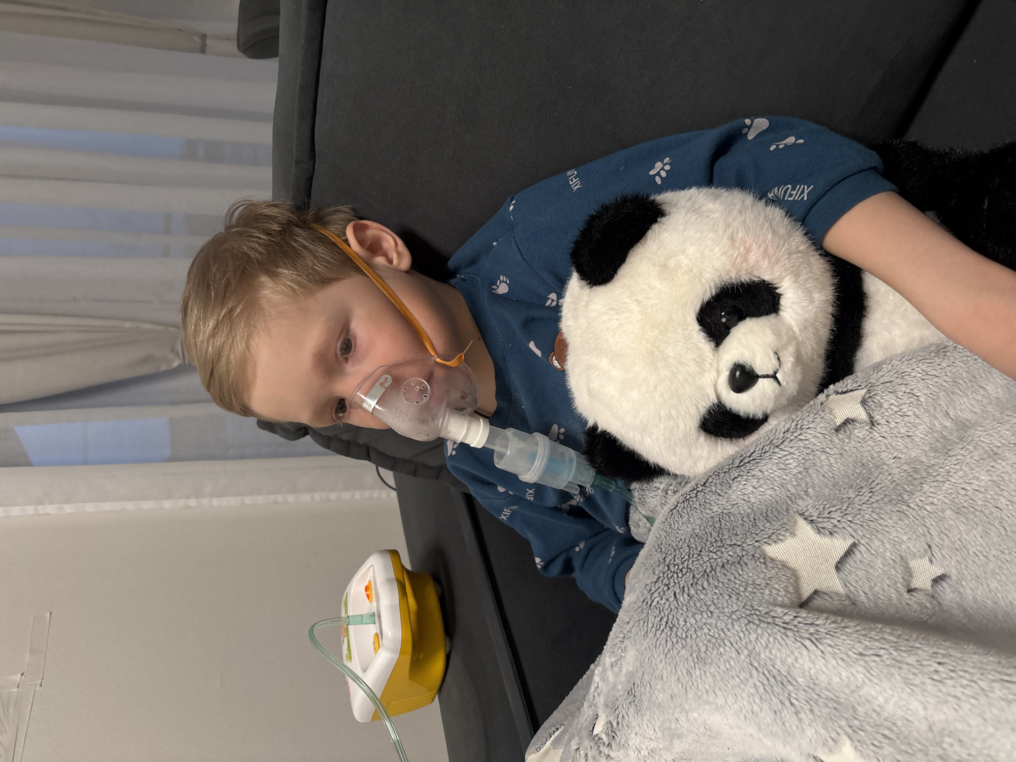 A young, light-haired boy is lying on a dark gray sofa, holding a stuffed panda toy. The boy is wearing a blue pajama top with white paw print designs. He has an orange-strapped nebulizer mask over his face. The nebulizer machine, which is yellow and white, sits on the floor next to the sofa. The boy and panda are partially covered by a gray blanket with white star patterns. The background shows a white wall and sheer white curtains. The location is not evident from the image.