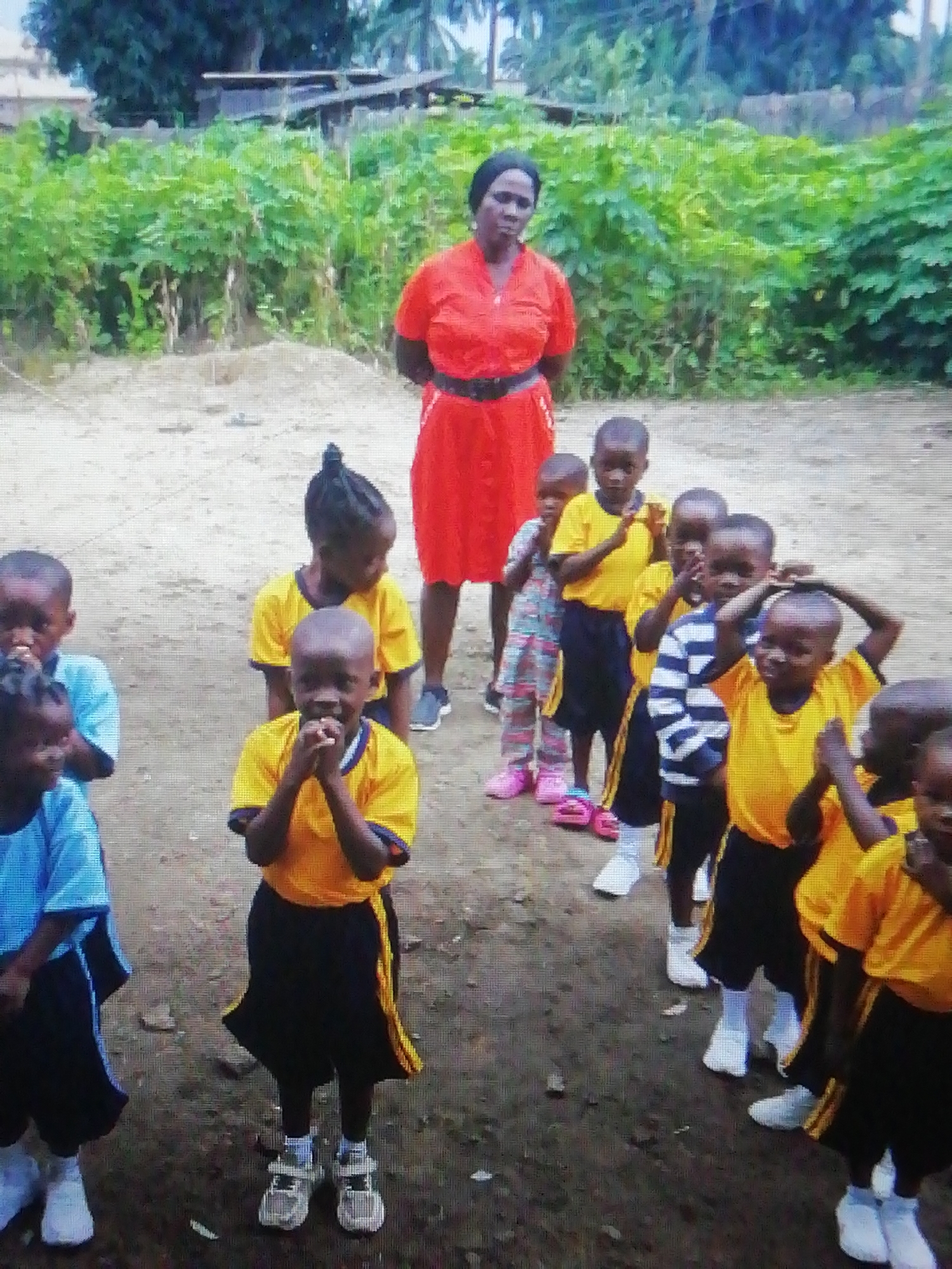 The image depicts a group of children and an adult, likely a teacher, in an outdoor setting. The children are dressed in school uniforms consisting of yellow shirts and black shorts with a yellow stripe. Some wear white socks and shoes or sandals. One child is wearing a blue shirt. One child is wearing a colorful outfit and pink sandals. The teacher is standing behind the children, wearing a red dress with a black belt. The background shows green vegetation, possibly bushes and trees. The ground appears to be dirt. The location of Enugu, Nigeria is not verifiable based on the provided image.