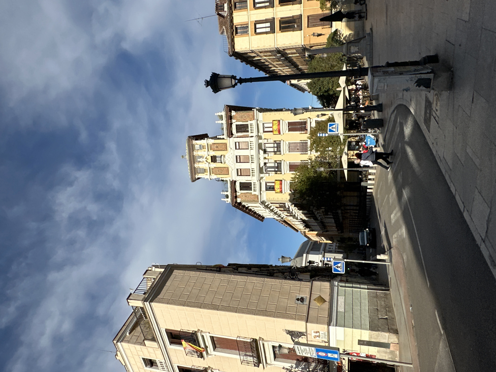 The image depicts a street scene in Madrid, Spain, characterized by a mix of architectural styles and urban elements. The buildings are a mix of beige and light tan colors.
People can be seen walking along the sidewalks and are sitting at outdoor cafe tables in the background.
Several elements define the street: streetlights, pedestrian crossing signs, and traffic signals. A vehicle is also visible on the street.
The sky is partly cloudy.