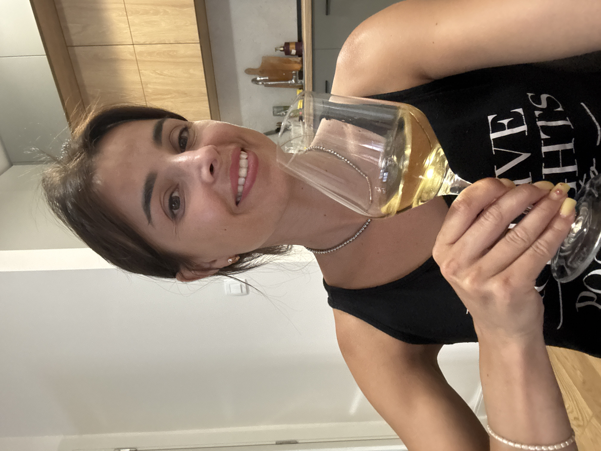 A woman is holding a glass of white wine and smiling at the camera. She is wearing a black tank top with white text on it and a pearl bracelet. The background appears to be a kitchen, with wooden cabinets and a sink visible. The lighting suggests it is daytime, likely indoors.