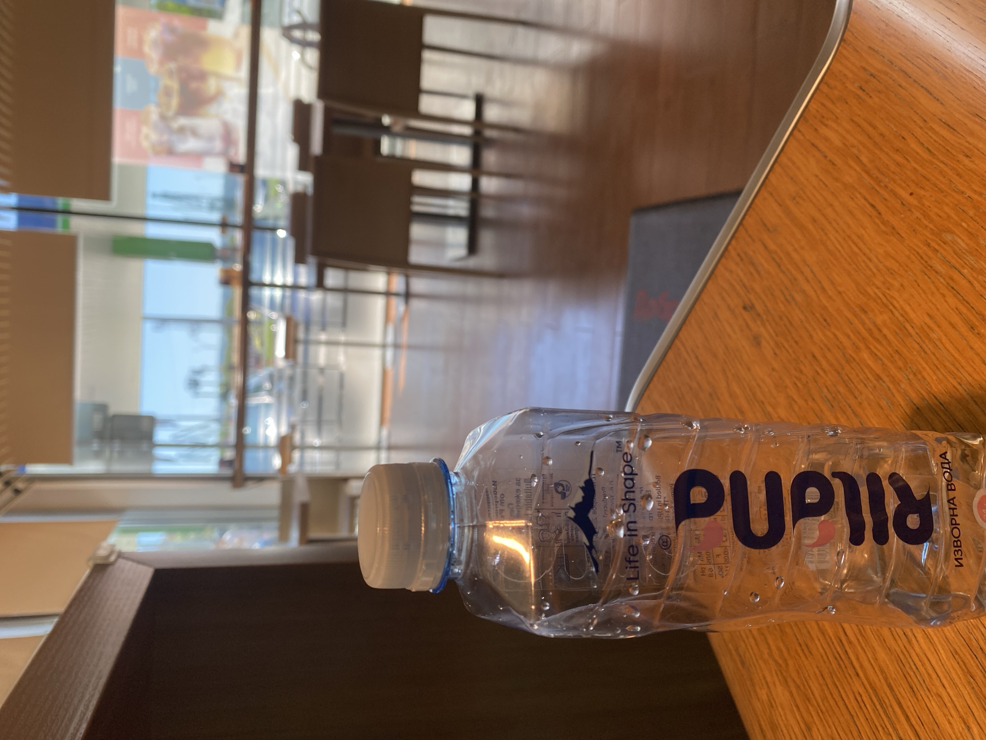 The image presents a close-up, eye-level shot of a partially consumed bottle of "Pupila" brand spring water, positioned on a wooden surface. The bottle features the words "Life in Shape TM" and "ИЗВОРНА ВОДА" in Cyrillic script, indicating that it is sourced from Bulgaria. The bottle's label also has the text: "39 - Every Day - 2017". The background reveals an interior setting that could be a cafe or a public space. Through a glass window, a glimpse of an outdoor scene is visible, featuring additional tables and chairs, implying an outside seating area. The location can be interpreted as being in the city of Varna, Bulgaria, based on the Cyrillic text on the water bottle.