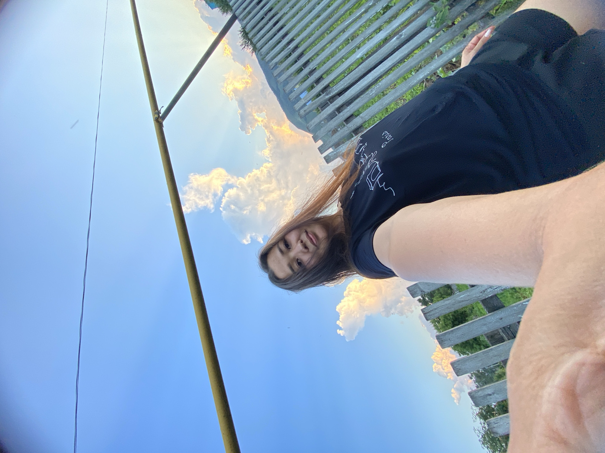 The image shows a woman in a black t-shirt and black shorts, taking a selfie outdoors. She is standing in front of a wooden fence, with a bright blue sky and clouds in the background. A yellow metal pipe runs across the top of the frame. The setting appears to be rural. Based on the context provided about Maloarslangulovo, Russia, this image was likely taken in the village of Maloarslangulovo.