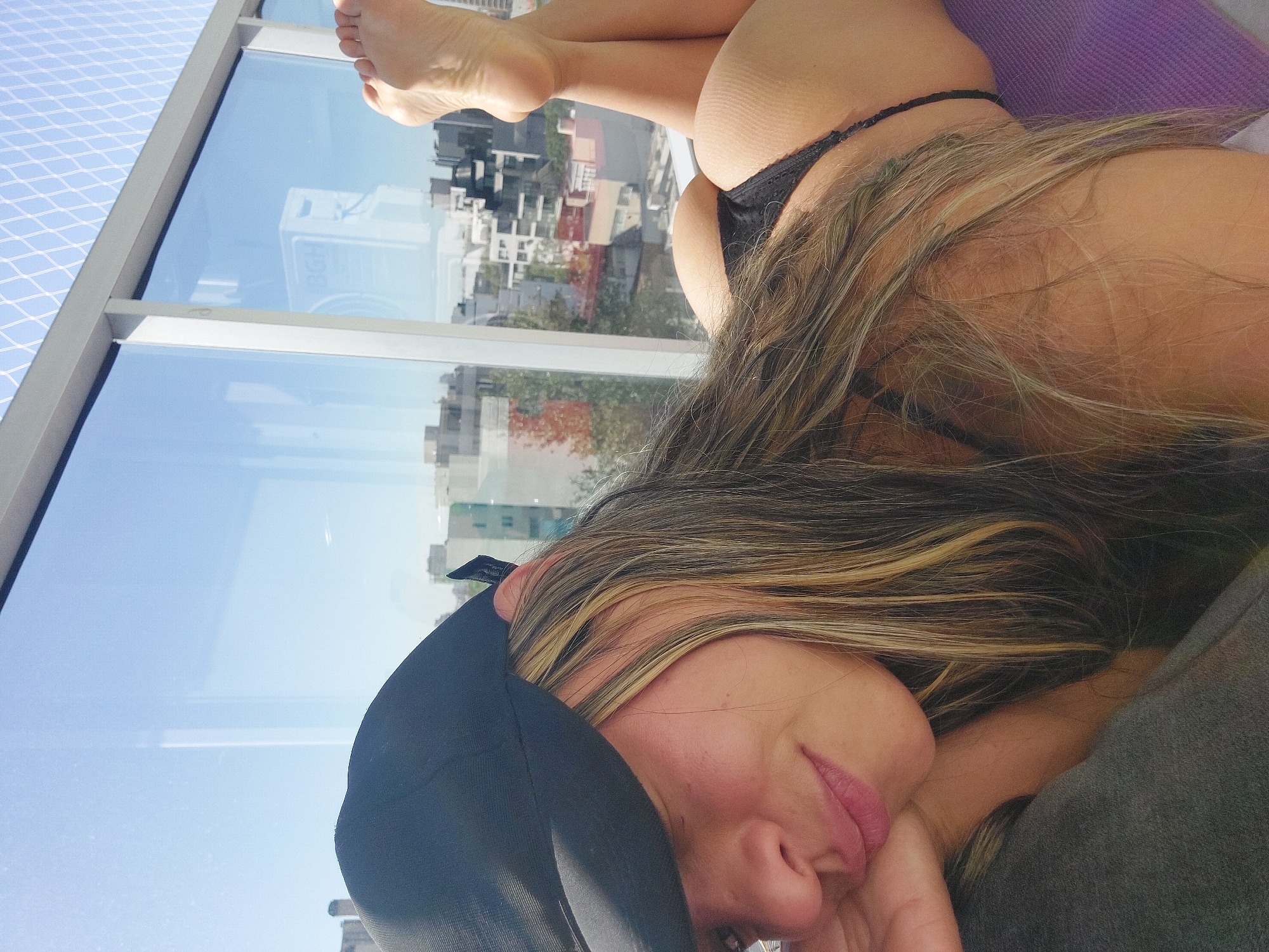A woman is depicted lying down indoors, next to a large window overlooking a city. Her body is positioned so that her head, shoulders, and lower body are visible. She is wearing a black baseball cap and a black thong. Her long, streaked blonde and brown hair flows over her shoulder and arm. Her face is partially visible, showing a relaxed expression with a subtle smile and pink lipstick.
Through the large glass window, a bright blue sky is reflected, along with urban buildings and trees. A white safety net with a grid pattern is visible outside the window frame on the left. The window's metal frame is also prominent. In the reflection on the window, the letters "BGH" are visible. The woman's bare feet and legs are visible near the top of the frame, and her buttocks are prominent in the black thong. She appears to be resting on a purple cushion or surface, with a grey fabric element near her head. The scene suggests a daytime setting in a high-rise building, consistent with an urban environment like Buenos Aires, Argentina.