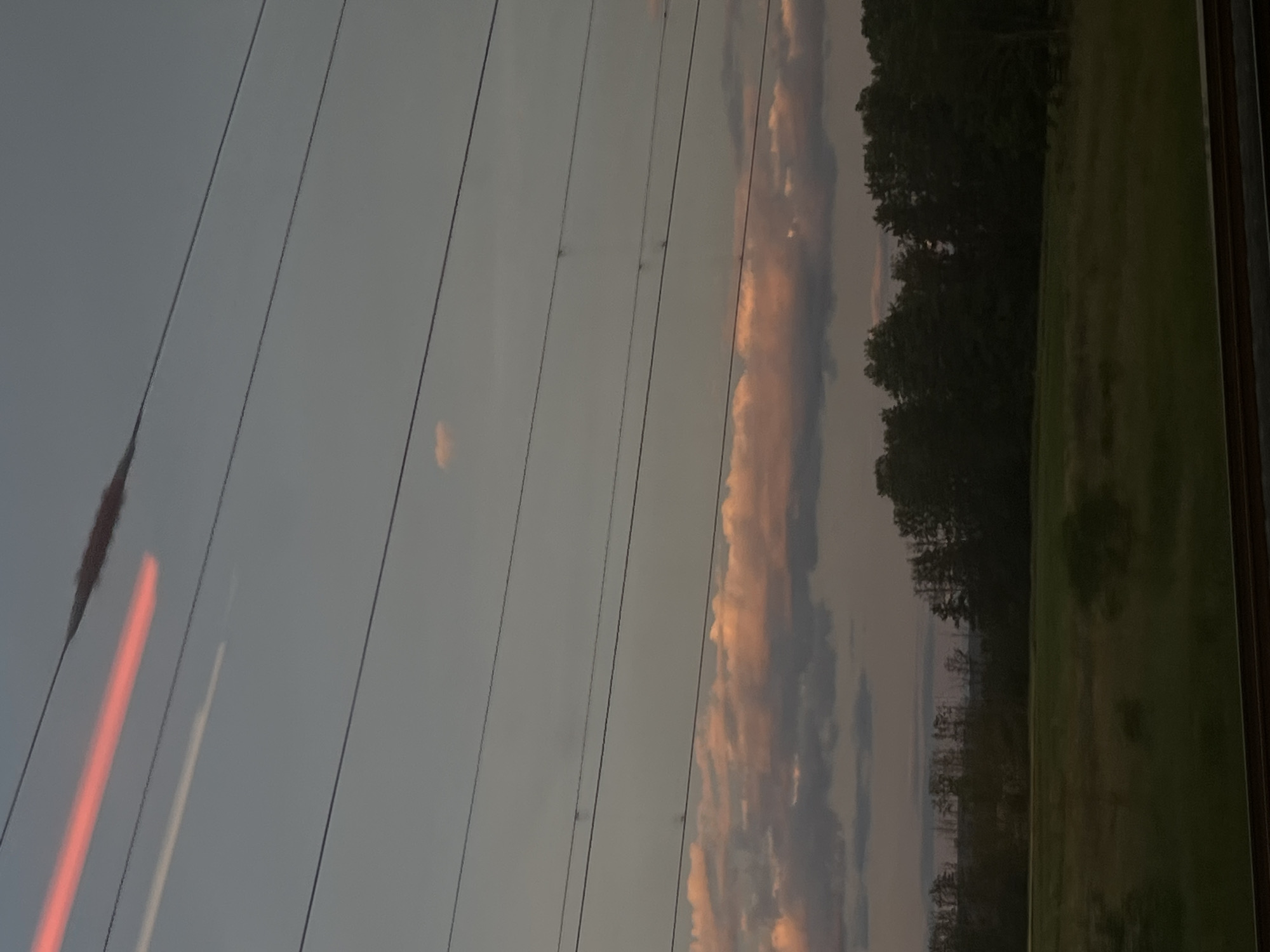 The image depicts a landscape scene with a dusky sky, illuminated by the soft glow of sunset. Several power lines stretch across the frame, dividing the sky into horizontal segments. In the upper portion of the sky, a blurred streak of bright red light is visible, possibly indicating motion blur from a light source or a lens flare. Below this, a faint white contrail or light streak is also present. The sky transitions to a paler grey towards the horizon.
A band of clouds, tinged with pink and orange hues from the setting sun, sits just above the treeline. These clouds appear textured and varied in density, with some darker grey clouds interspersed. The treeline is composed of dark silhouettes of numerous trees, creating a dense, somewhat indistinct barrier. The land in front of the trees is a field of grass, appearing somewhat muted in color due to the fading light. In the foreground, a glimpse of what appears to be a railroad track or similar structure with dark horizontal beams is visible at the very bottom of the image. The overall atmosphere is serene and tranquil, characteristic of twilight. The scene is devoid of any visible people or discernible human activity.