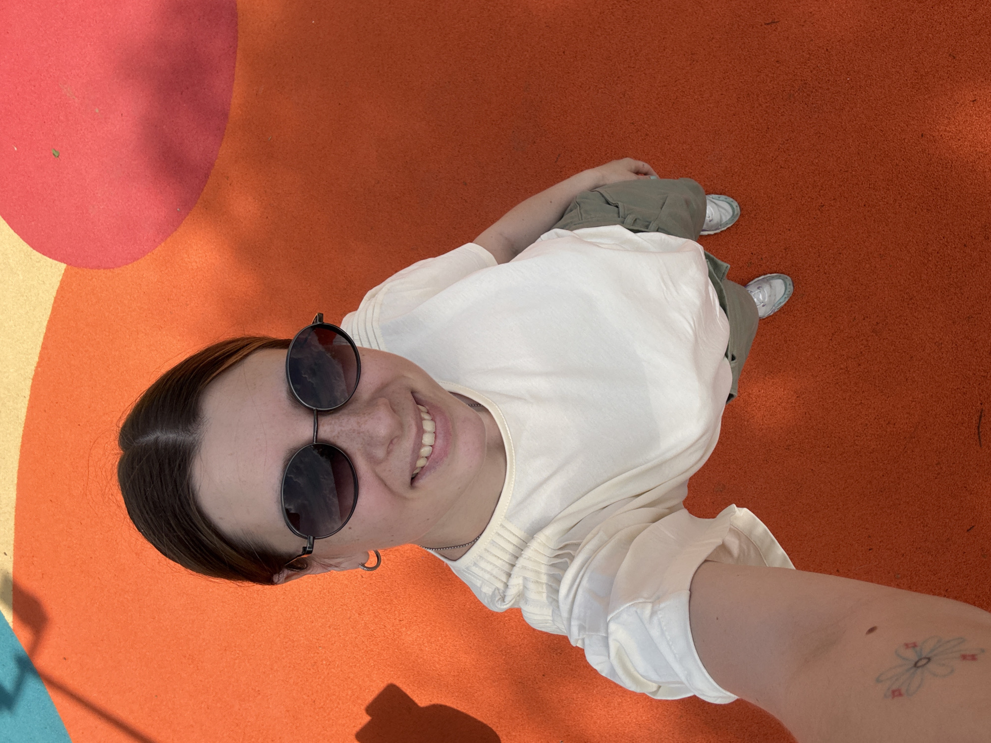 A selfie taken from a high angle features a young woman with dark hair pulled back, wearing round sunglasses, a white long-sleeved shirt, and green pants, standing on a surface with orange and red sections. She is smiling at the camera. Her right arm is visible with a flower tattoo on the forearm. The location appears to be an outdoor public area in the city of Moscow, Russia.