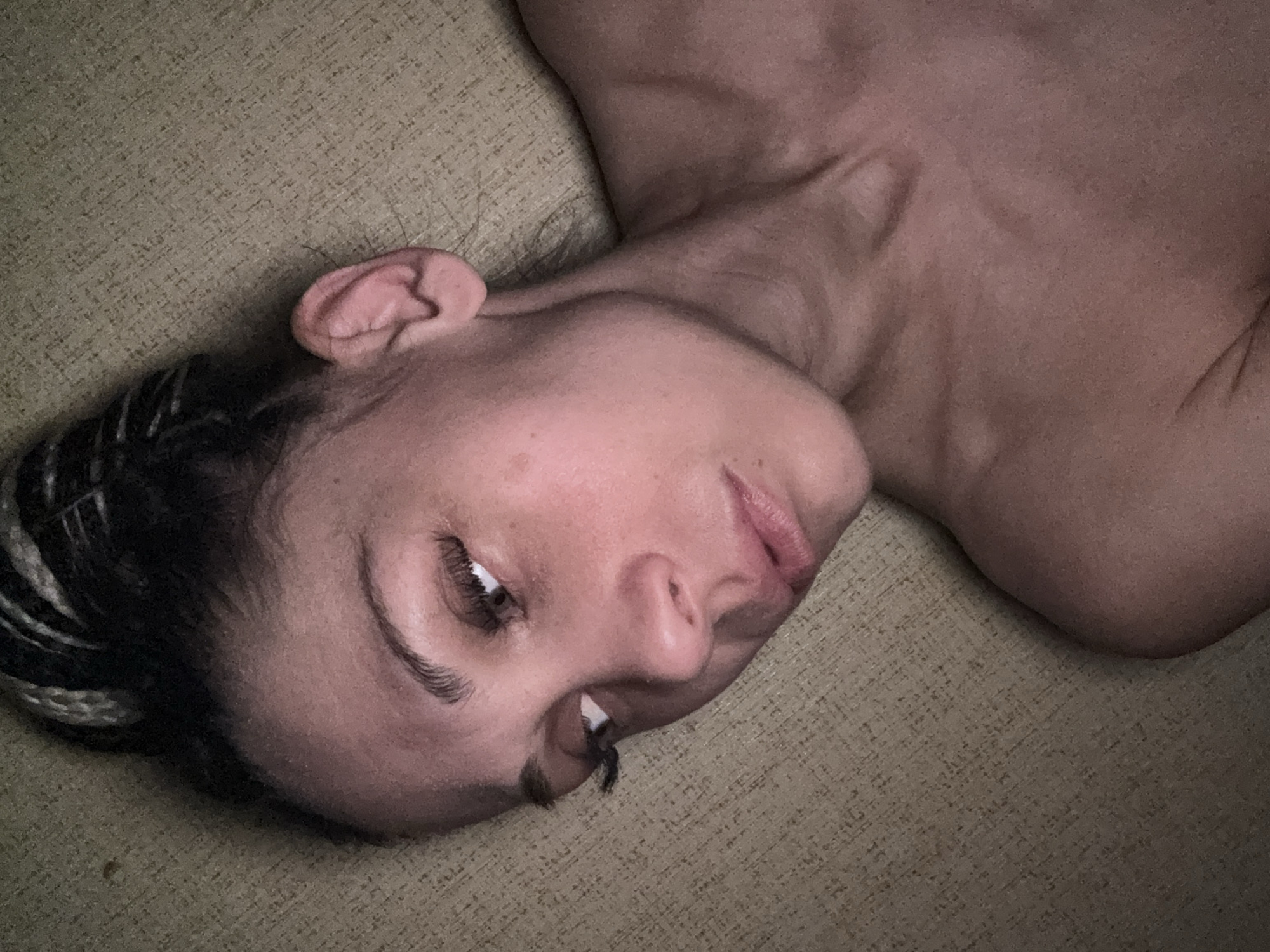 A person with light skin is lying down on a textured surface. The person has braids in their hair and is looking towards the left of the image. The person's head and neck are visible, as is a portion of their shoulder. The background is out of focus, but appears to be a fabric-covered surface. The location in the city is unknown.