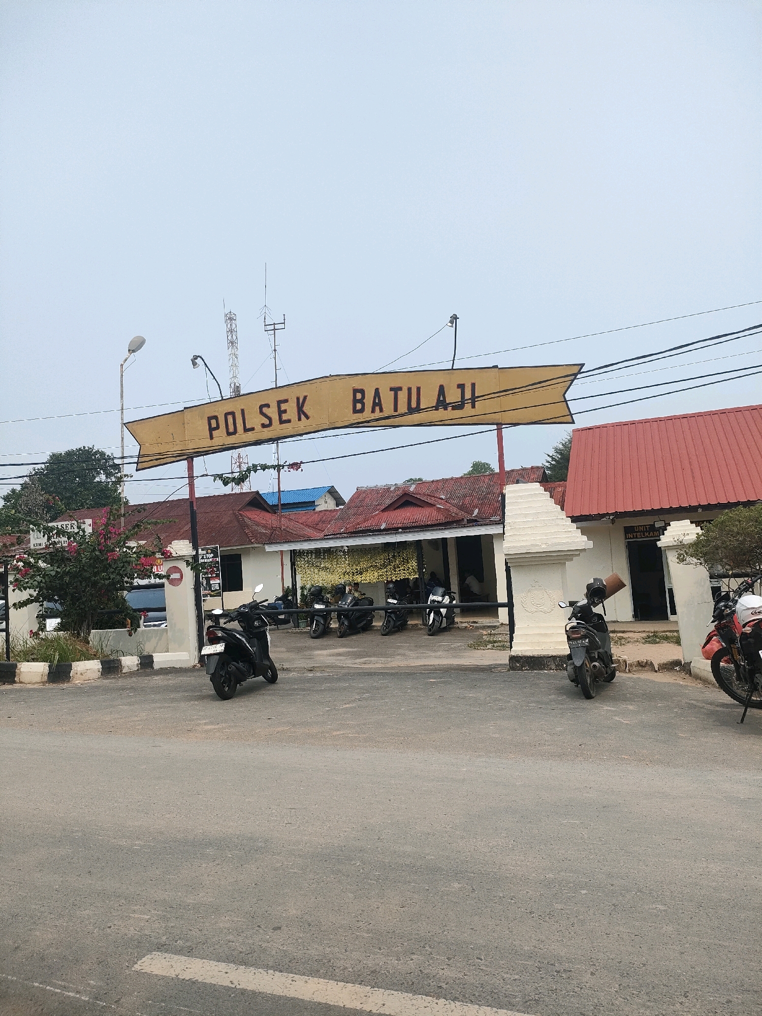 The image shows the entrance to the POLSEK BATU AJI police station in Batam, Indonesia. A yellow sign with black lettering hangs above the entrance, reading "POLSEK BATU AJI". The building appears to be a single-story structure with a red tiled roof. Several motorcycles are parked in front of the building. A white pillar with ornate carvings stands to the right of the entrance. The road in front of the police station is asphalt, and there are some plants and trees in the foreground. The sky is overcast.