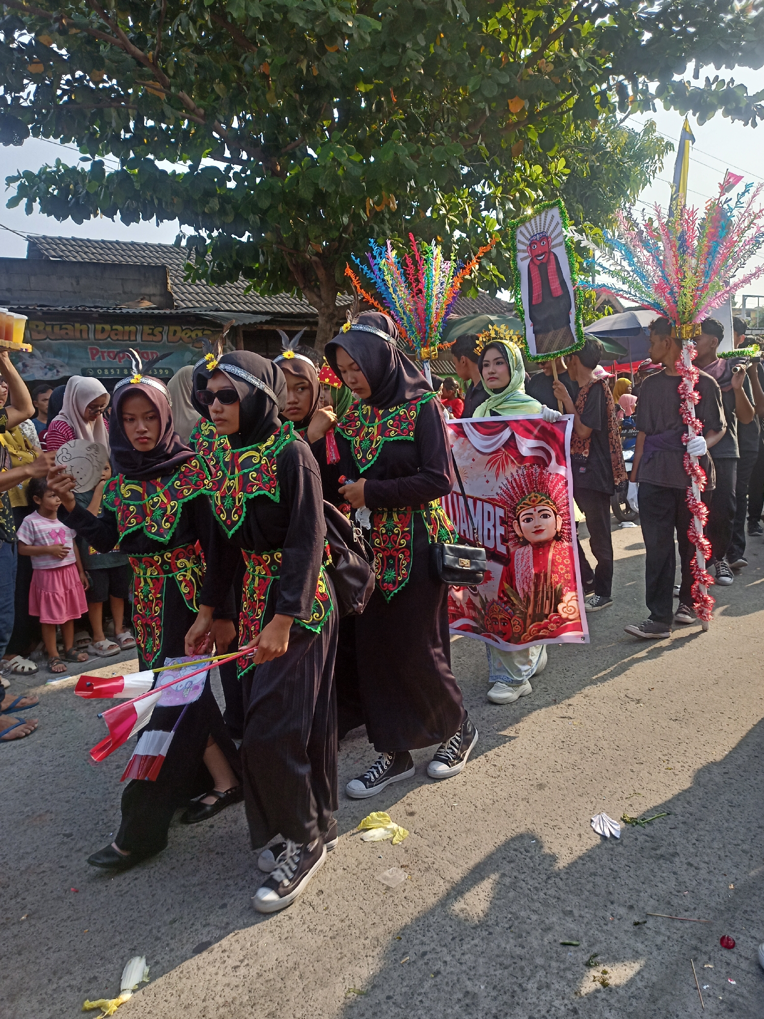 In Saren, Indonesia, a group of people are participating in a parade on a sunny day. In the foreground, four young women in matching black outfits with colorful embroidered collars are walking. The woman on the far left is holding a small circular object with an image of a face. The women are wearing headscarves and some have decorative headpieces. Behind them, other individuals are also in the procession. One person is carrying a large banner featuring a red puppet-like figure with traditional attire and text. Another individual is holding a pole adorned with red and white trim. In the background, there are trees, buildings, and a blue and yellow flag. The parade is taking place on a paved road with debris scattered on the ground, suggesting a festive or celebratory event.
