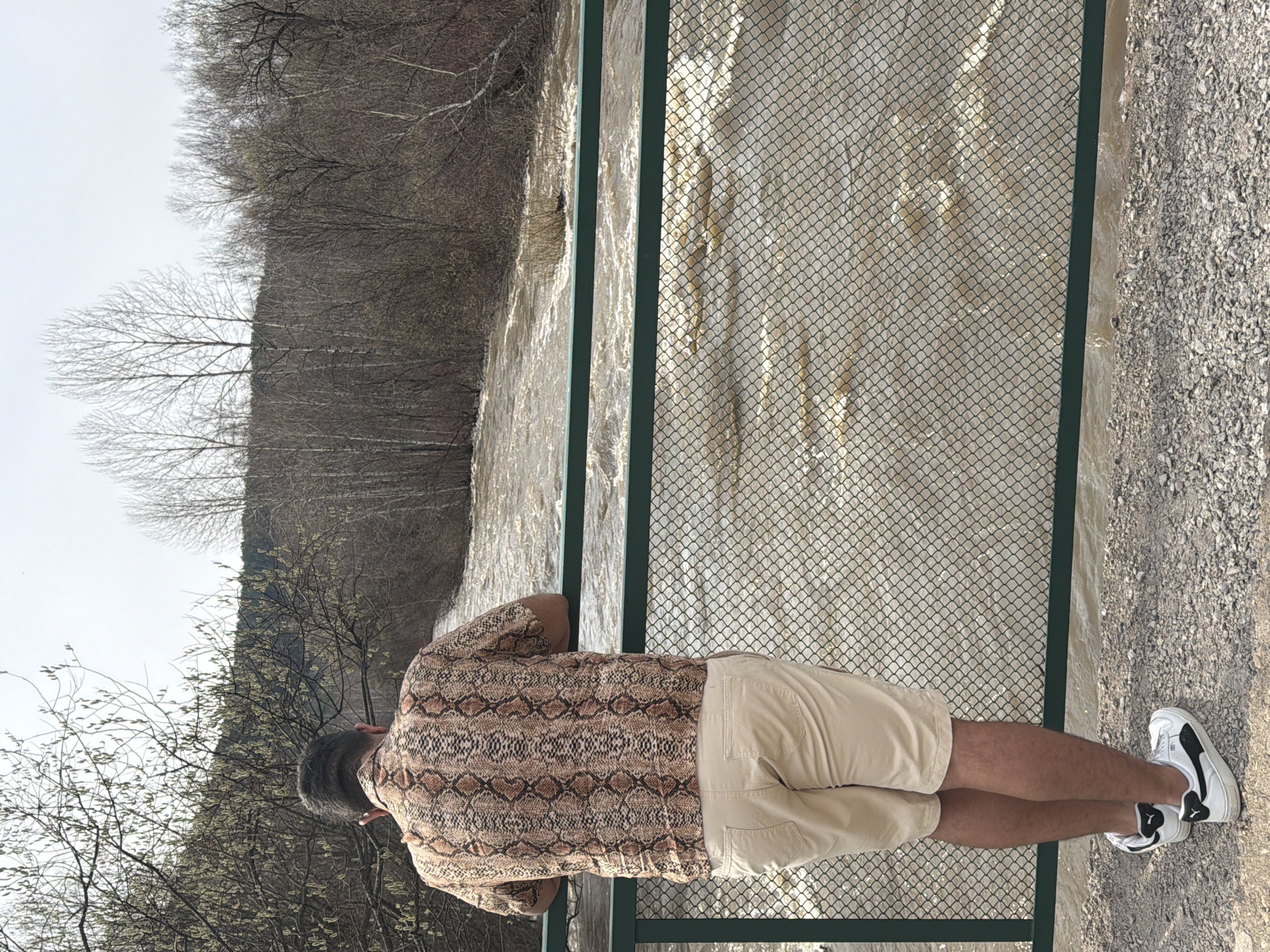 A man with dark hair, wearing a snakeskin-patterned shirt, tan shorts, and white sneakers, stands on a gravelly surface, leaning slightly against a green metal fence. Beyond the fence is a body of water, likely a river, with murky, fast-flowing water. On the far bank of the river, bare trees form a dense forest. The sky is overcast. The location is likely in Krasnoyarsk, Russia, though no definitive markers confirm this information. The man appears to be observing the river.
