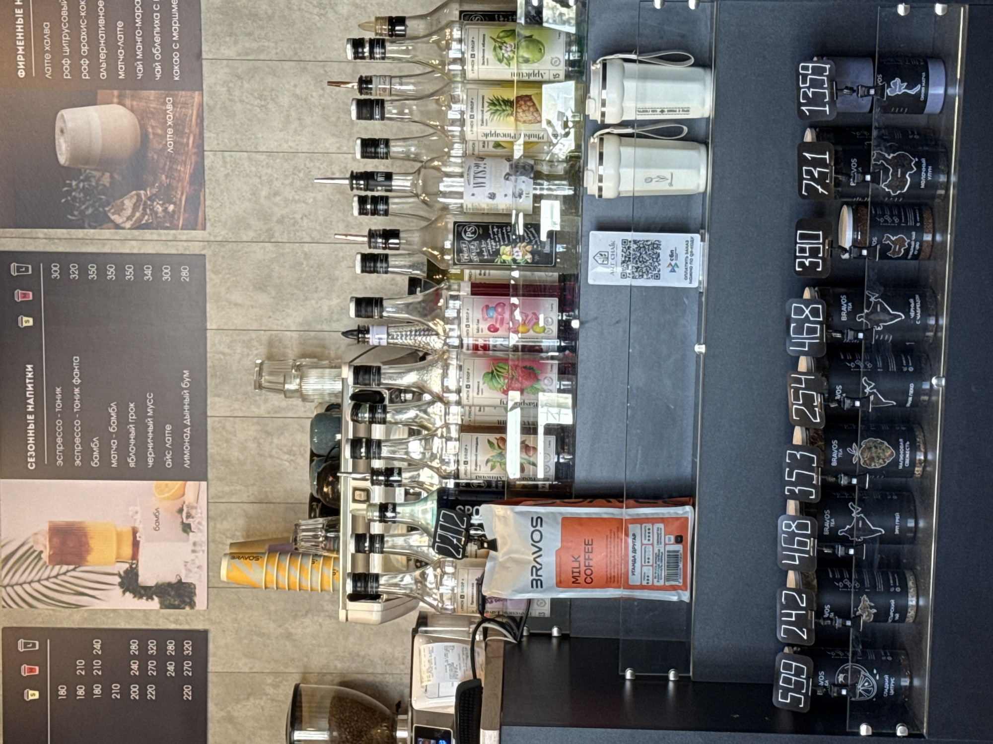 This image captures a well-organized service counter, likely in a cafe or coffee shop in Chaikovsky, Russia. The scene displays various ingredients and products for sale, including syrups, tea, and coffee.
**Subjects and Objects:** The central focus is a transparent display case containing numerous clear glass bottles, each fitted with a pour spout and labeled with different flavorings like "Appletini," "Phuket Pineapple," "Raspberry," and "Almond." Below these bottles, a coffee grinder with visible beans stands next to small yellow and orange "BRAVOS" paper cups. Adjacent to the syrups is a retail display of black cylindrical containers of tea from "BRAVOS TEA," each with a white handwritten price. Examples include "СЛАДКИЙ ЦИТРУС" (Sweet Citrus) priced at 599, "ЭРЛ ГРЕЙ" (Earl Grey) at 468, "МАЛИНОВАЯ СВЕЖЕСТЬ" (Raspberry Freshness) at 353, "ЧЕРНЫЙ С ЧАБРЕЦОМ" (Black with Thyme) at 468, "МОЛОЧНЫЙ УЛУН" (Milk Oolong) at 390 and 731, and "СИНЯЯ МАТЧА" (Blue Matcha) at 1359. A bag of "BRAVOS MILK COFFEE" named "УГАНДА ДРУГАР" (Uganda Другар) is also visible.
**Activity and Event:** The arrangement suggests a commercial setting for preparing and selling beverages. Customers would select drinks from the displayed menus and potentially purchase the packaged tea. The QR code indicates a modern payment method.
**Notable Details:** The image includes two menu boards listing "ФИРМЕННЫЕ НАПИТКИ" (Signature Drinks) and "СЕЗОННЫЕ НАПИТКИ" (Seasonal Drinks) with their prices. Signature drinks include "латте халва" (latte halva) at 300 and "раф цитрусовый" (citrus raf) at 320. Seasonal drinks list "эспрессо - тоник" (espresso tonic) and "матча - бамбл" (matcha bumble). A QR code labeled "ART CHAIK" with text "ОПЛАТИТЬ ЗАКАЗ можно по QR-коду" (Pay for order can be done by QR code) and "сбп" (SBP - System of Fast Payments) is prominently displayed. The overall impression is one of efficiency and product variety.