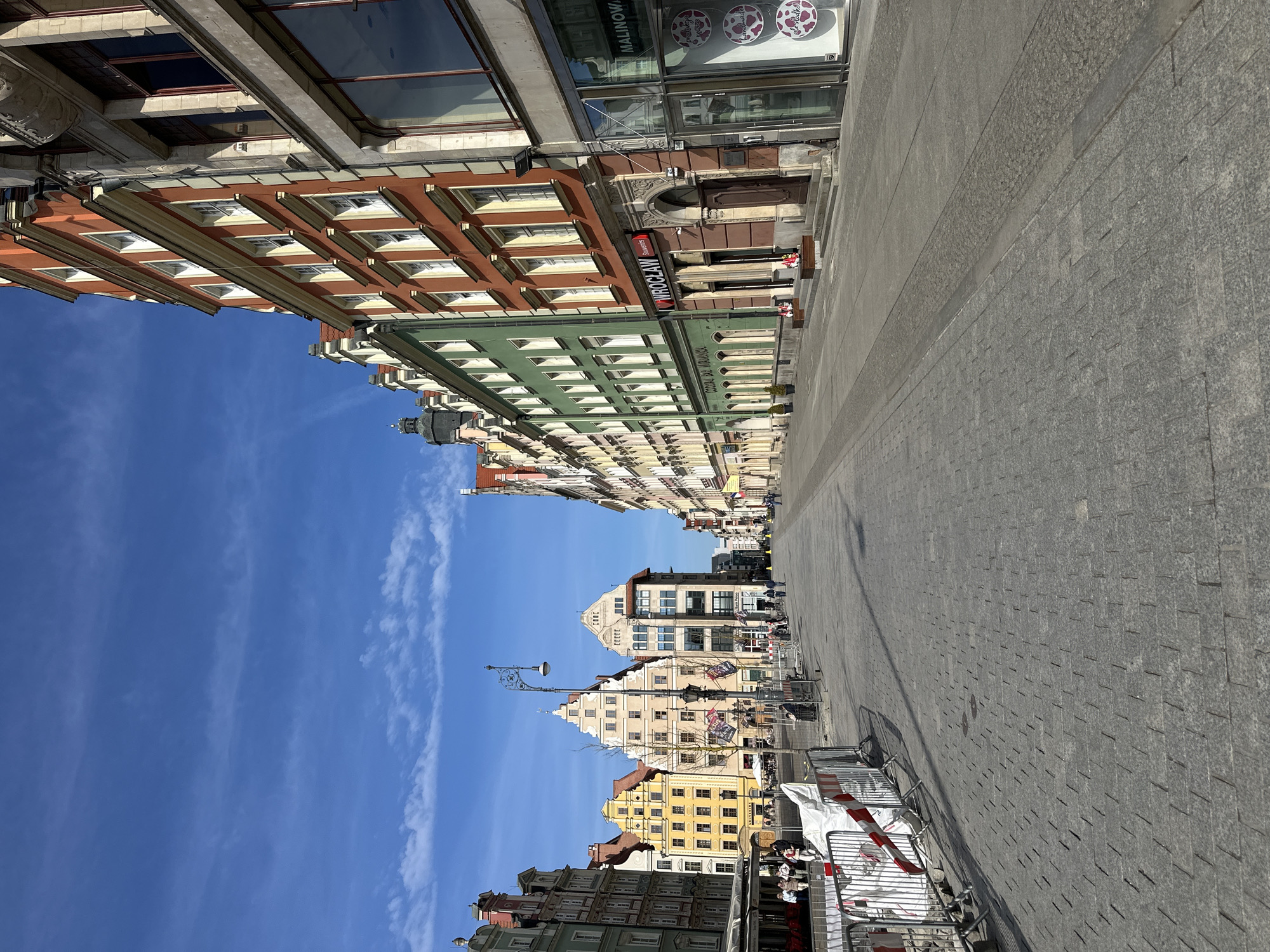The image captures a street view in Wrocław, Poland, characterized by historical buildings with varied architectural styles and facades. The sky is a clear blue with faint clouds. A pedestrian zone paved with stone dominates the foreground, lined by metal barriers and construction supplies. Buildings are visible on both sides of the street, featuring multiple stories and windows. The buildings' exteriors vary in color from brown and red to green and yellow, showcasing diverse designs and ornamentation. A street lamp is standing close to the middle part of the buildings. Signage on the buildings is visible, including one that reads "WROCŁAW Souvenirs". People are visible in the background, appearing as small figures due to the distance. The perspective suggests the photograph was taken from street level.