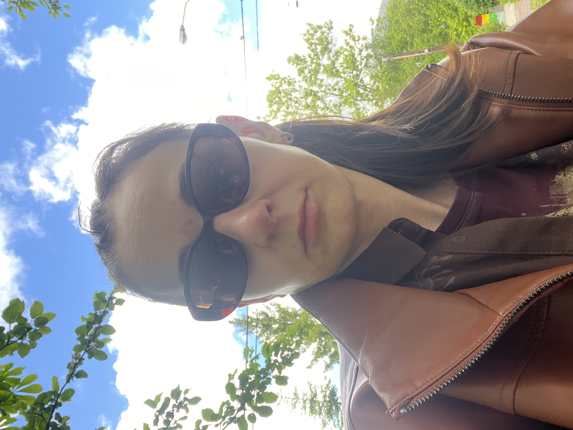 A woman wearing dark sunglasses and a brown leather jacket is visible. Her hair is dark brown and styled down. She is outdoors, with a background of blue sky with white clouds and green trees. The location appears to be an urban environment, suggested by a lamppost and what appears to be a building in the distance.