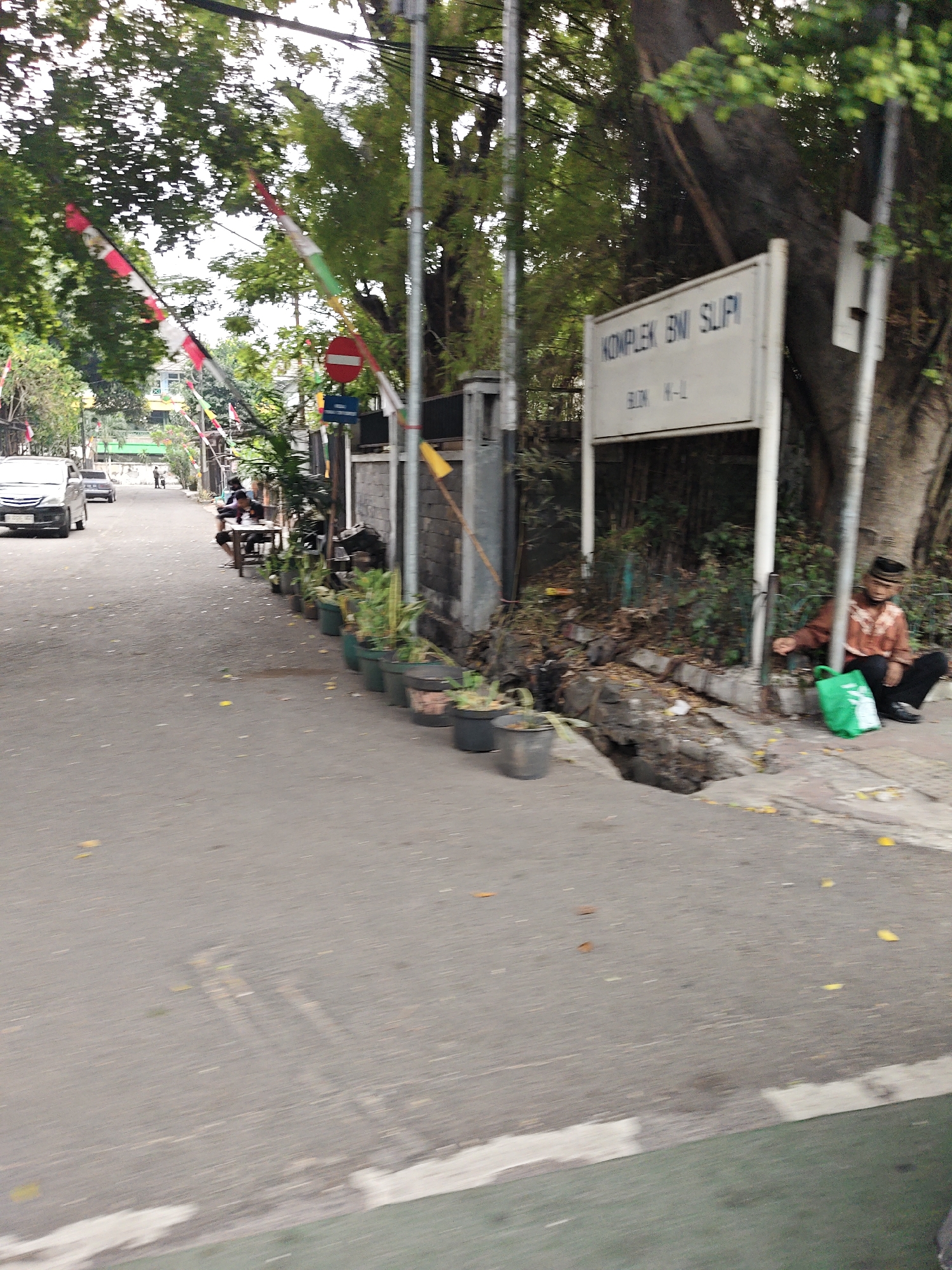 The image depicts a street scene in the city of West Jakarta, Indonesia. A white sign with blue text reads "KOMPLEKS BNI SLIPPI BLOK H-L". A person is sitting on the side of the road next to a large tree, wearing a reddish-brown shirt and black pants, with a black skullcap. A bright green bag is on the ground next to them. There are several potted plants lining the street. A white van is parked further down the road, and other cars are visible in the distance. Red and white flags are strung along the street, suggesting a festive or commemorative occasion. The road surface appears to be asphalt, with some debris and fallen leaves scattered about. The overall impression is that of a residential or commercial street in an urban environment.