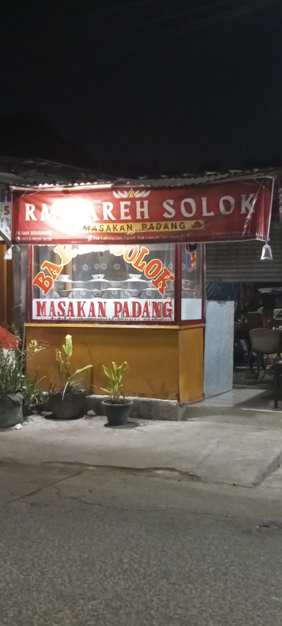 The image shows a roadside restaurant in Setu, Indonesia, called "RM AREH SOLOK". The restaurant has a prominent red banner with white text that reads "RM AREH SOLOK" and "MASAKAN PADANG". Below the banner, on the glass storefront, are the words "BAKUL OLOK" in large, stylized red letters. Inside the store, several bowls are neatly arranged on shelves. The restaurant is lit by artificial lights, and the surroundings suggest it is nighttime. In front of the restaurant, there are two potted plants on the concrete sidewalk. The overall scene depicts a typical Indonesian street food establishment.