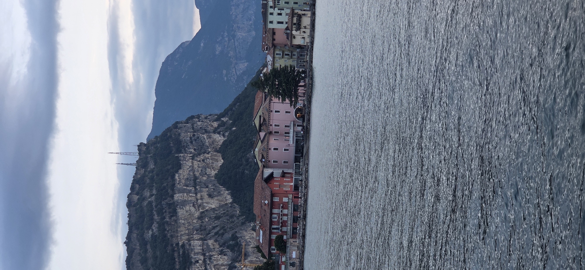 The image captures a waterfront scene in Torbole, Italy, on an overcast day. In the foreground, the rippling surface of a body of water occupies the lower half of the frame. Beyond the water, a row of buildings lines the shore. The architecture is varied, with several multi-story structures featuring pitched roofs. Some buildings are painted in warm tones like red and pink, while others are lighter in color, such as pale yellow and green. A large, dark green evergreen tree stands prominently among the buildings.
Behind the town, a massive, steep, rocky cliff face dominates the background, its surface textured and weathered. The upper edges of the cliff are covered with dark green vegetation. Two tall, lattice-like radio towers are visible on the crest of the cliff, silhouetted against the cloudy sky. Further in the distance, the soft outlines of more mountains recede into the horizon. The sky is a muted grey with patches of lighter clouds. No people are visible in the scene. The overall impression is one of a tranquil, natural landscape meeting a settled, historical town.