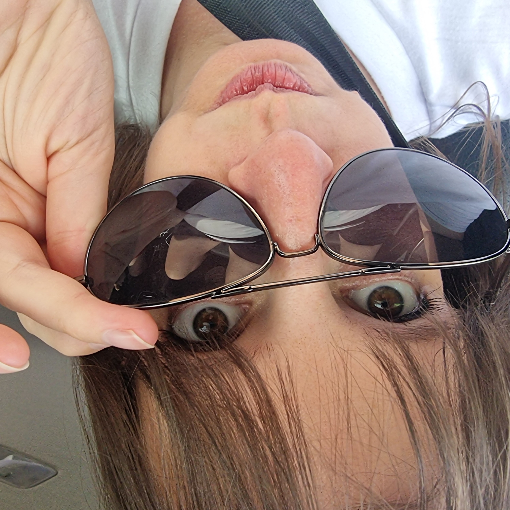 In Dunnae-myeon, South Korea, a close-up, frontal selfie features a person looking directly at the camera. The individual is wearing a white t-shirt and sunglasses, which they are adjusting with their right hand. The lighting suggests an outdoor environment, possibly a car, as indicated by the reflections in the sunglasses. The person's hair, which is brown and falls around their face, is slightly tousled. The image is focused on the upper portion of the person's face, including their eyes, nose, and mouth, with the sunglasses obscuring a significant portion of their eyes. The background is indistinct, hinting at a seated position.