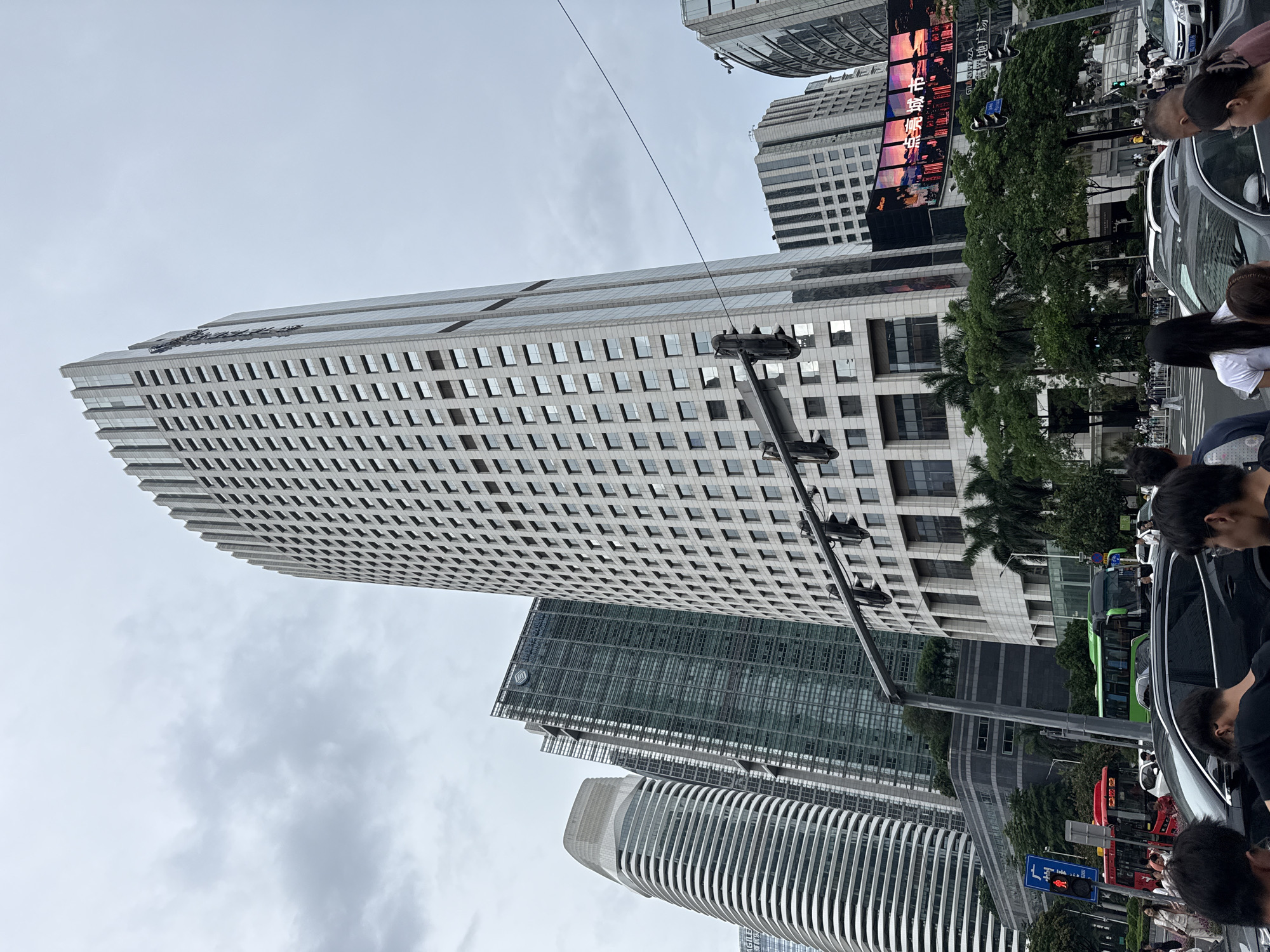 In Tianhe District, China, a city scene unfolds under an overcast sky. The focal point is a tall, modern skyscraper with a light-colored facade, its rectangular windows forming a grid pattern. To the left and right of this central building are other contemporary skyscrapers, including one with a curved, ribbed exterior and another clad in dark glass. In the foreground, traffic signals hang over a busy intersection. Several cars and buses, including a red bus and a green bus, are visible on the road. Pedestrians are crossing the street at a designated crosswalk, while others stand on the sidewalk. A large digital billboard displays a vibrant image of a city skyline at sunset. Lush green trees and palm trees dot the landscape, providing a contrast to the urban architecture. The overall impression is one of a dynamic and bustling urban environment.