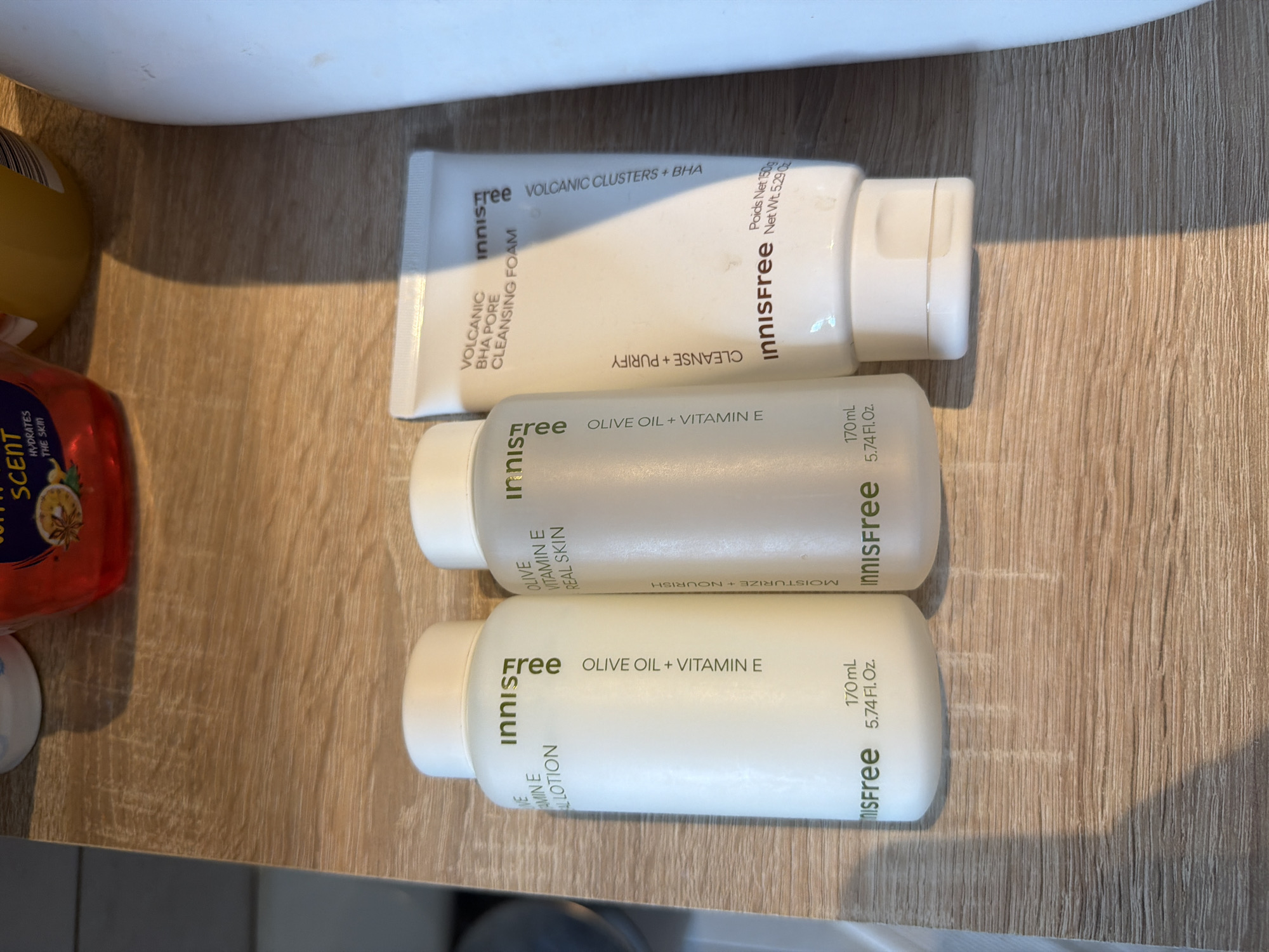The image shows three white tubes/bottles of Innisfree skincare products lying on a wooden surface. The top product is "VOLCANIC BHA PORE CLEANSING FOAM", the middle one is "OLIVE OIL + VITAMIN E REAL SKIN", and the bottom one is "OLIVE OIL + VITAMIN E ALLOTION". A bottle of "AUTUMN SCENT" with an orange-red liquid inside is partially visible on the left side. The background appears to be a light-colored wooden surface with linear patterns. The location is impossible to determine based on the image.
