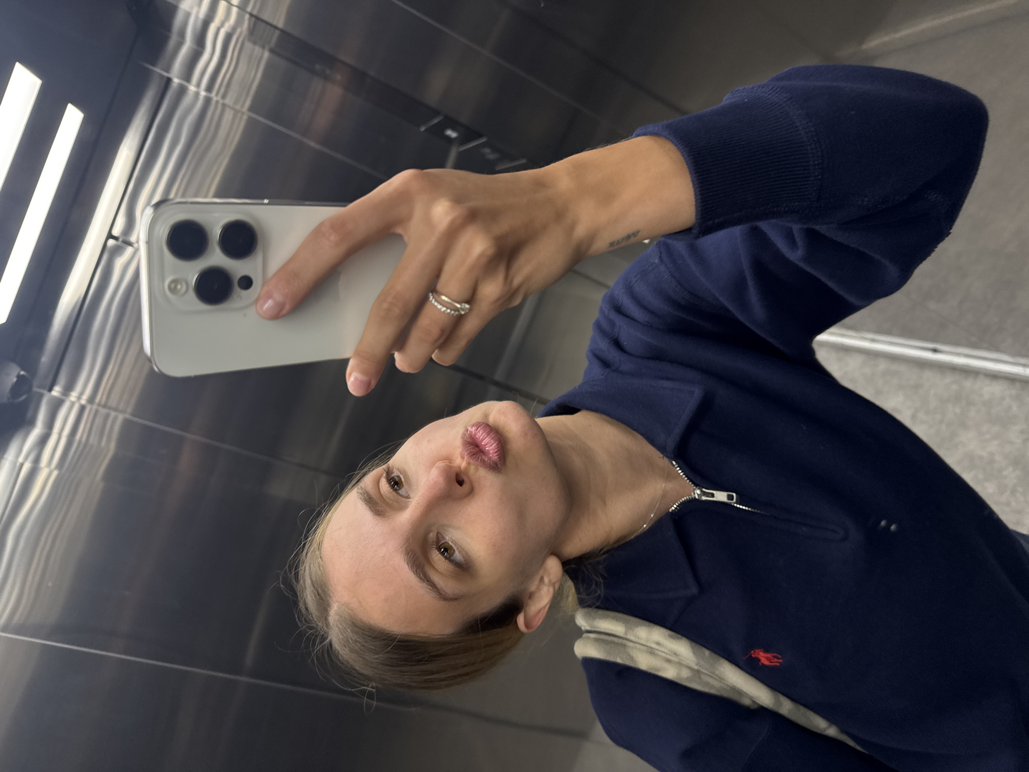 A woman is taking a selfie in an elevator. She is holding a white iPhone with multiple camera lenses. The woman has fair skin, brown eyes, and blonde hair pulled back. She is wearing a navy blue zip-up polo shirt with a small red logo. Her lips are pursed. She has a ring on her finger and a small tattoo on her wrist. The elevator has metal walls and ceiling with fluorescent lighting. The location in the city is Unknown, Unknown.