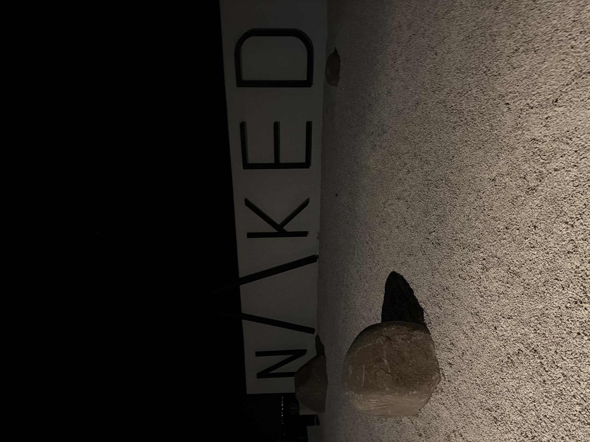 A nighttime, eye-level, medium shot captures the exterior of a building featuring the word "NAKED" in large, black, block letters mounted on a white facade. The letters are spaced out, with the "N" appearing slightly separated from the rest of the word. The ground in front of the building is covered in light-colored gravel, and three large, rounded rocks are positioned on the gravel. One rock is in the left foreground, partially casting a shadow. Another rock is to the left of the building's facade, and a third, smaller rock is to the right, near the "D" of "NAKED". The background is dark, with the faint outlines of lights suggesting a distant city or shoreline. The image has a somber and minimalist aesthetic. This scene is located at the entrance to the NAKED Beach in Burgas, Bulgaria.